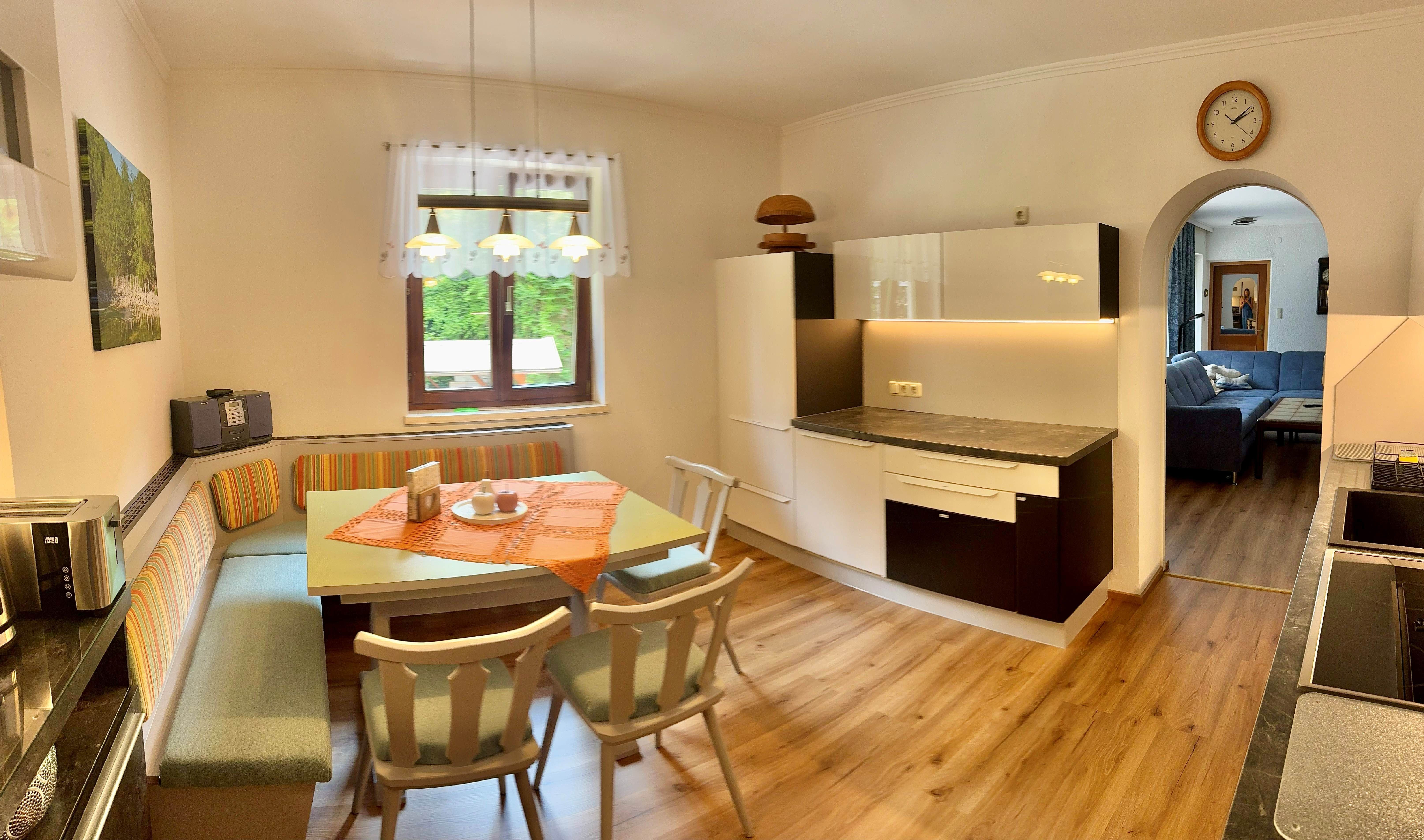 Gemütliche Küche mit Esstisch und Eckbank, daneben moderne Küchenschränke. Im Hintergrund ein Wohnzimmer mit Sofa.