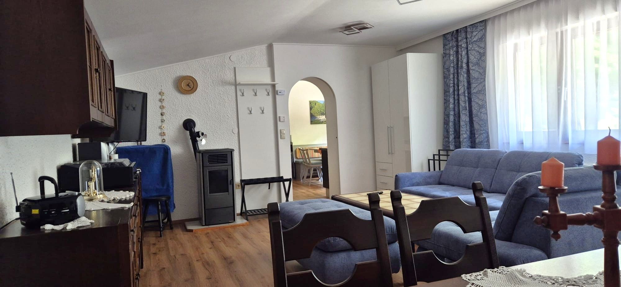 Gemütliches Wohnzimmer einer Ferienwohnung mit blauen Sofas, Holzmöbeln und einem Kamin. Ein Esstisch ist im Hintergrund sichtbar.