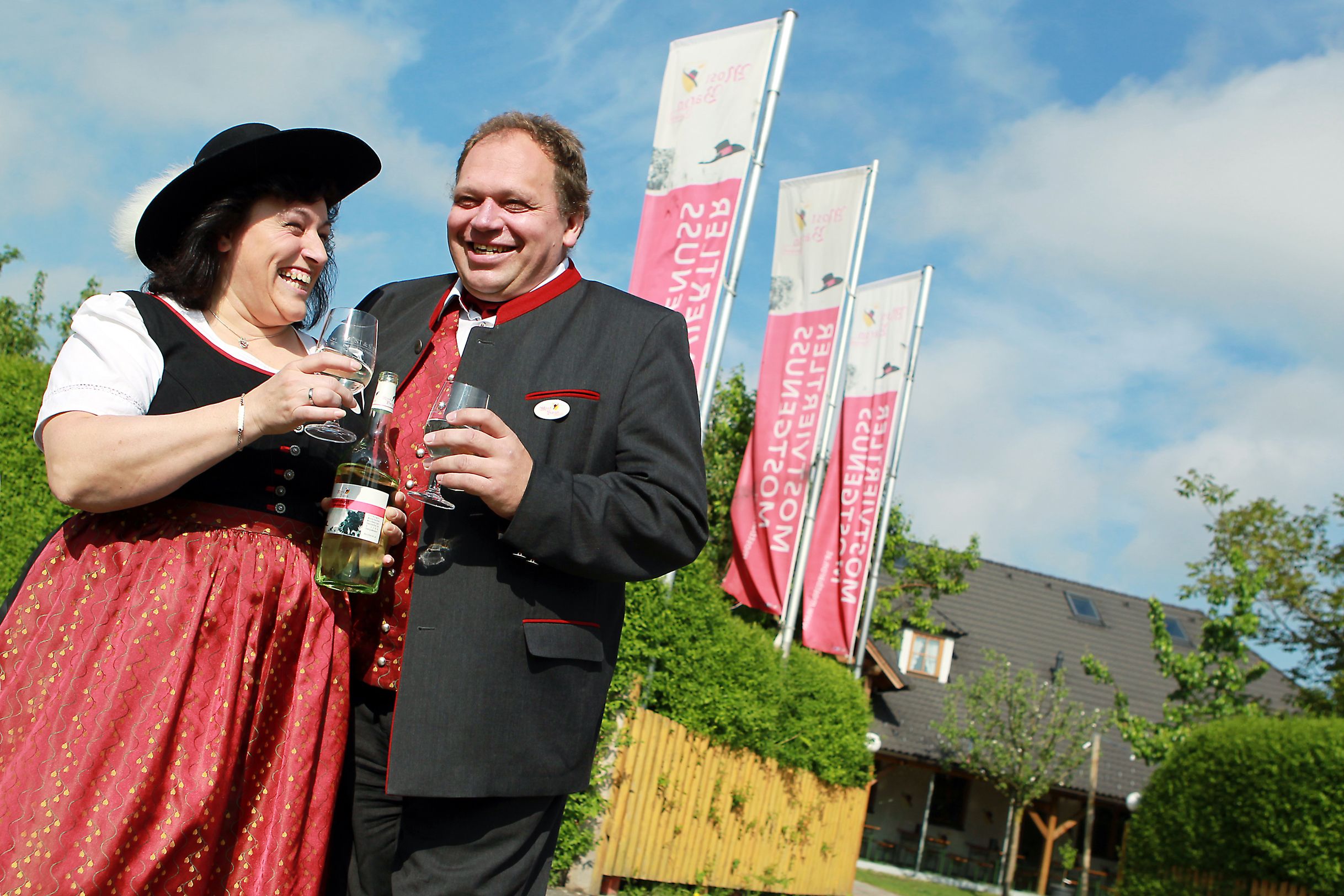 Ein Mann und eine Frau in traditioneller Kleidung stoßen mit Weingläsern an, im Hintergrund sind Fahnen und ein Gebäude zu sehen.