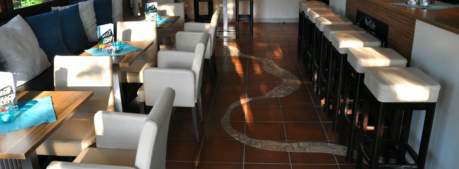 Innenansicht eines Cafés mit weißen Stühlen und Tischen, braunem Fliesenboden und Barhockern.