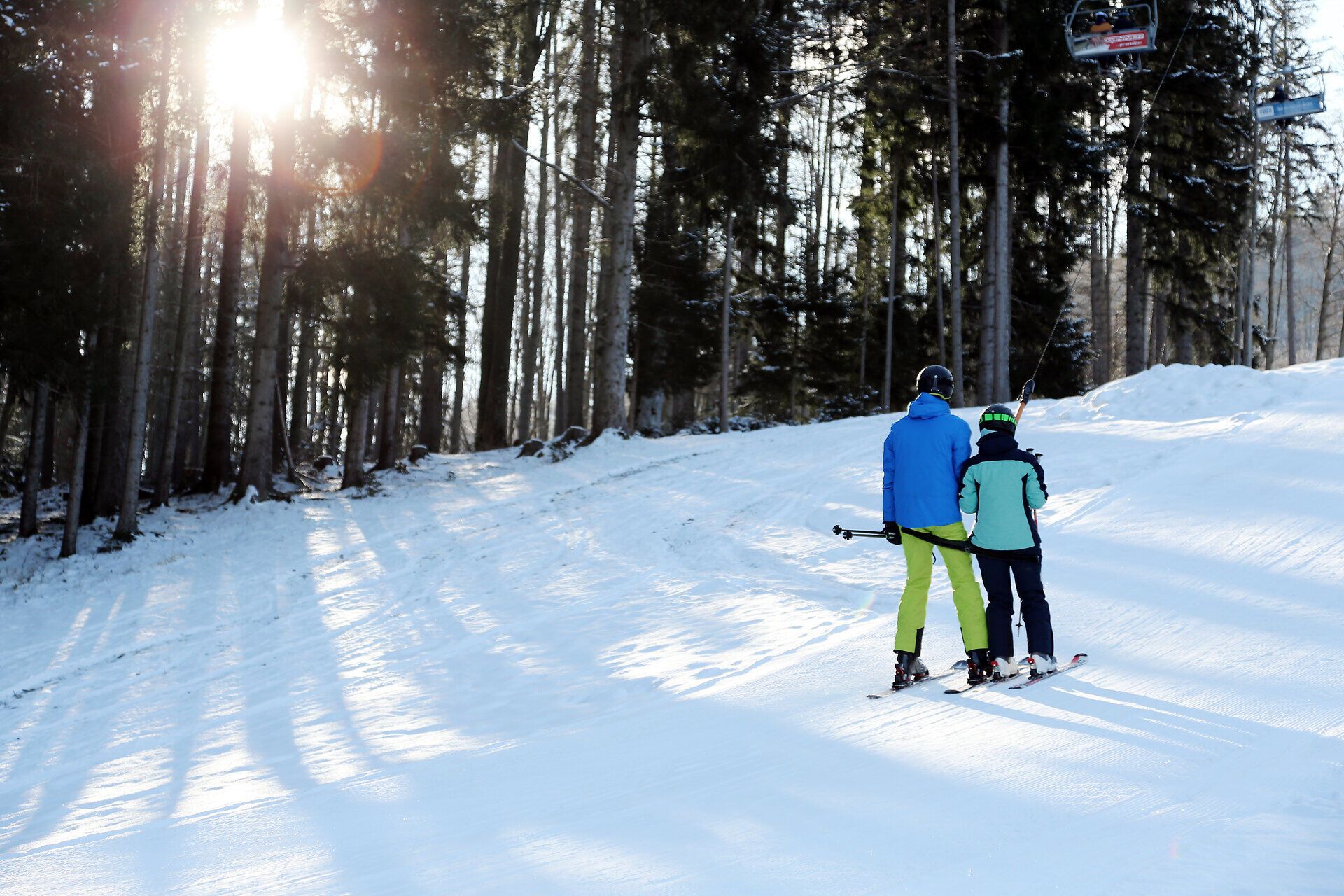 Eine Familie genießt den Wintertag auf der Forsteralm, während sie sich auf den Weg zum Schlepplift macht. Die schneebedeckten Bäume und die strahlende Sonne schaffen eine zauberhafte Atmosphäre, die zum Skifahren einlädt.