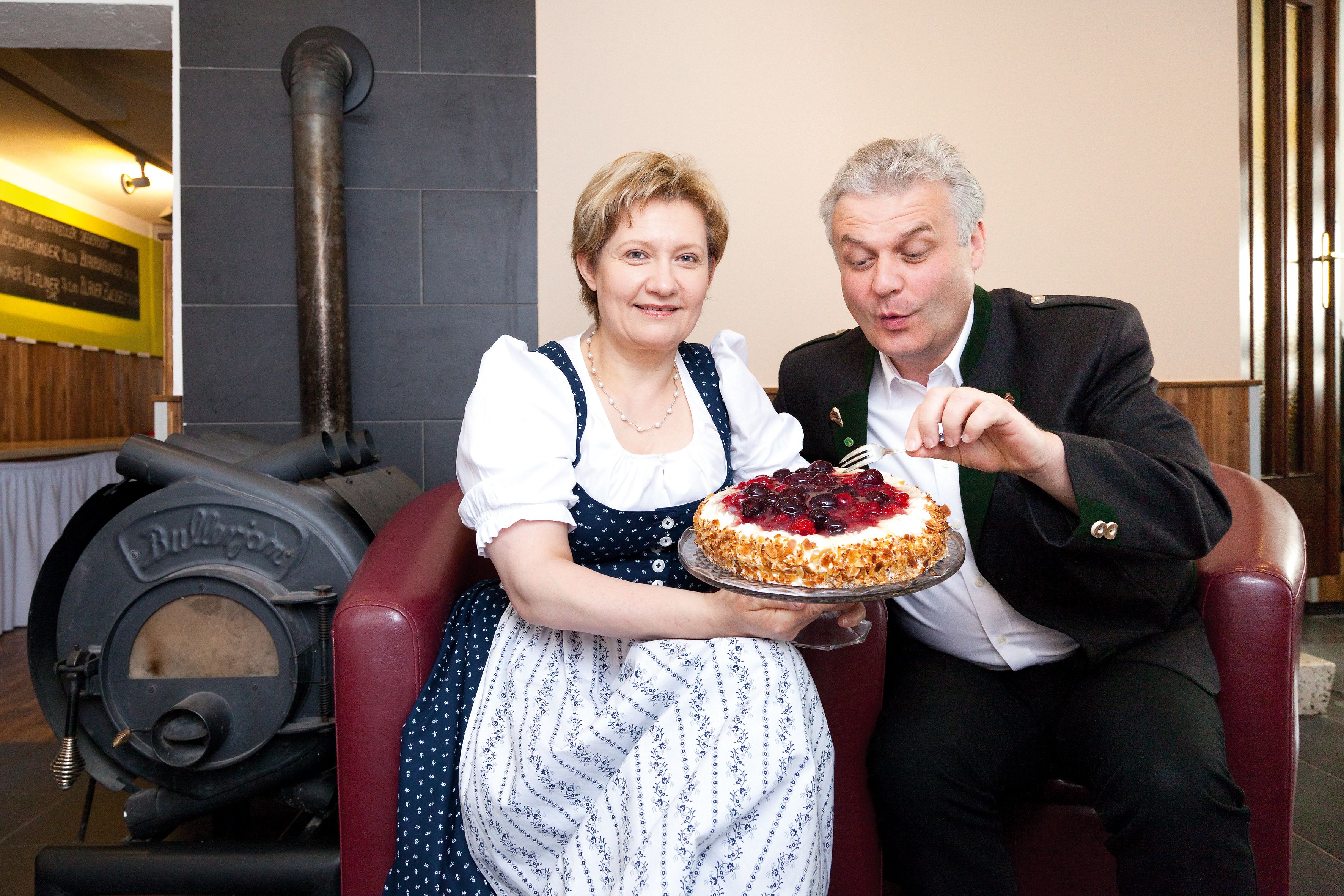 Ein Mann und eine Frau in traditioneller Kleidung halten eine Torte vor einem Ofen.
