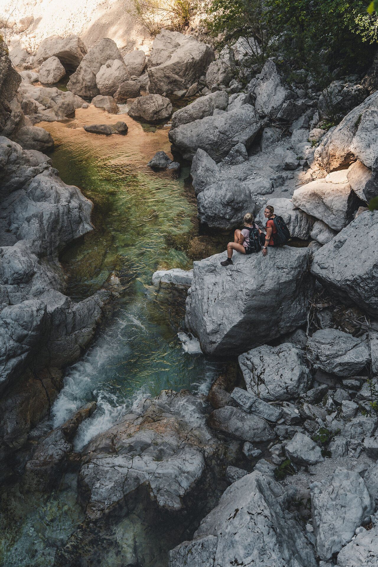 Zwei Frauen sitzen auf großen Felsen an einem klaren Fluss, umgeben von natürlicher Vegetation