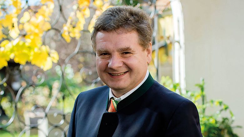 Ein lächelnder Mann in traditioneller Kleidung steht im Freien vor einem schmiedeeisernen Zaun mit gelben Blättern im Hintergrund.