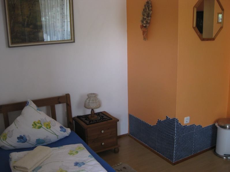 Ein kleines Zimmer mit einem Einzelbett, Nachttisch, Lampe und orangefarbener Wand mit Mosaikfliesen.