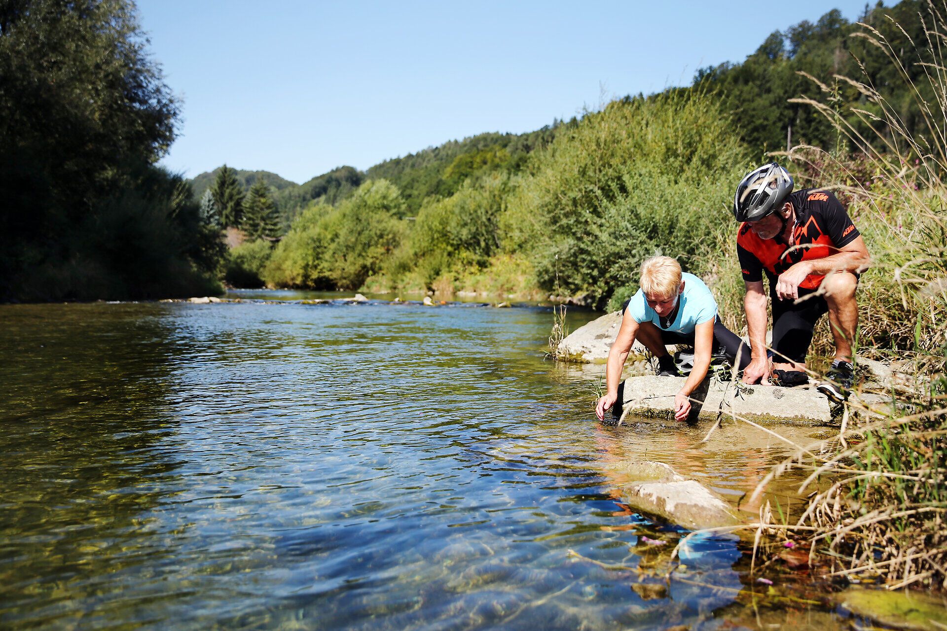 An einem sonnigen Tag am Ufer eines klaren Flusses erkunden zwei Abenteurer die Natur. Die sanften Hügel im Hintergrund und das glitzernde Wasser schaffen eine friedliche Atmosphäre, die zum Verweilen einlädt. Hier, wo die Schönheit der Landschaft auf die Freude am Radfahren trifft, wird jeder Moment zum unvergesslichen Erlebnis.