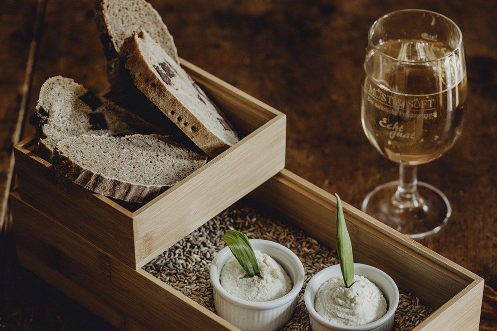 Ein Holztablett mit Brotscheiben, zwei Schälchen mit Aufstrich und ein Glas Weißwein auf einem Holztisch.