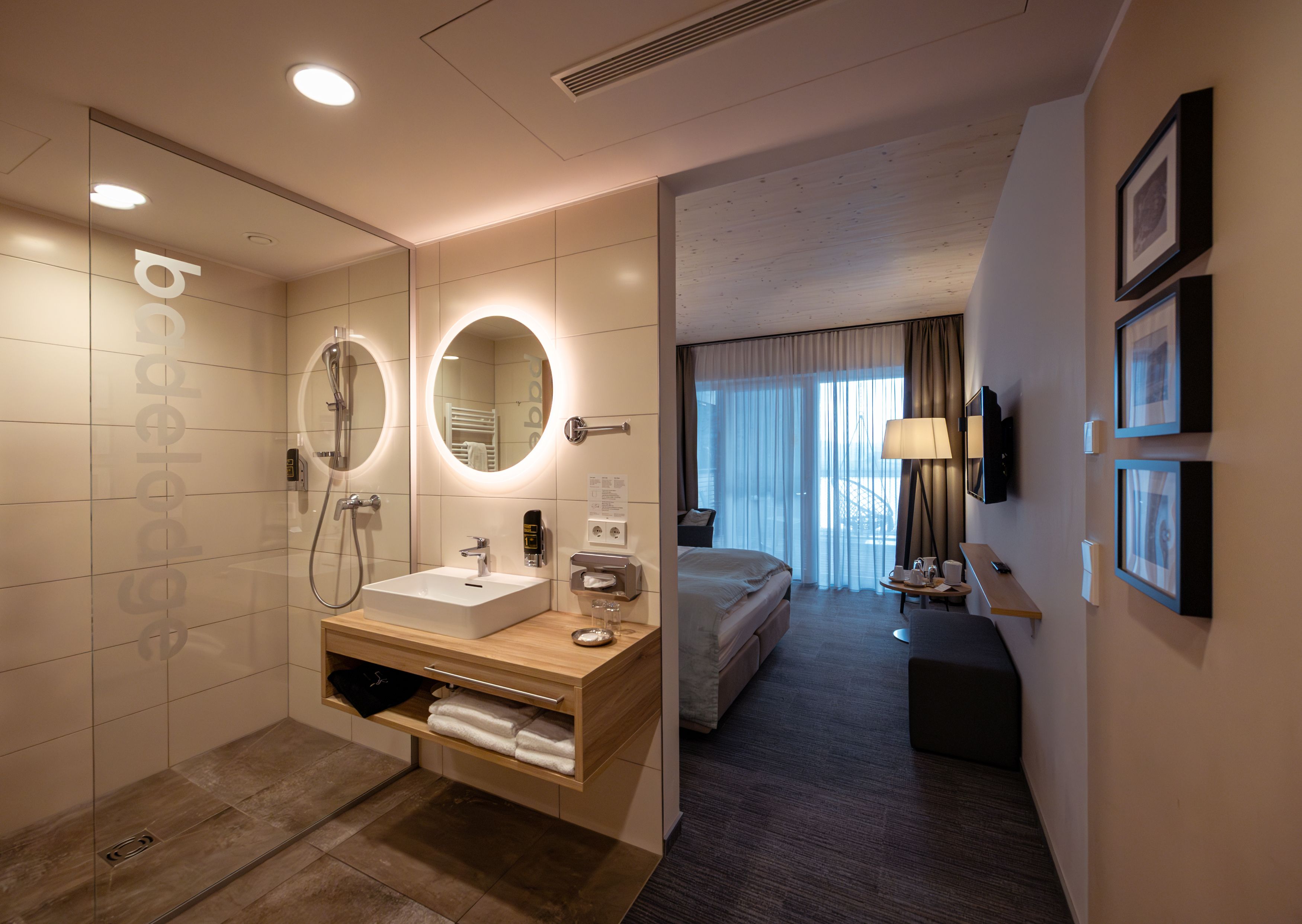 Modernes Hotelzimmer mit offenem Bad, Waschbecken, Spiegel und Dusche. Im Hintergrund ein Bett und ein Fenster mit Vorhängen.