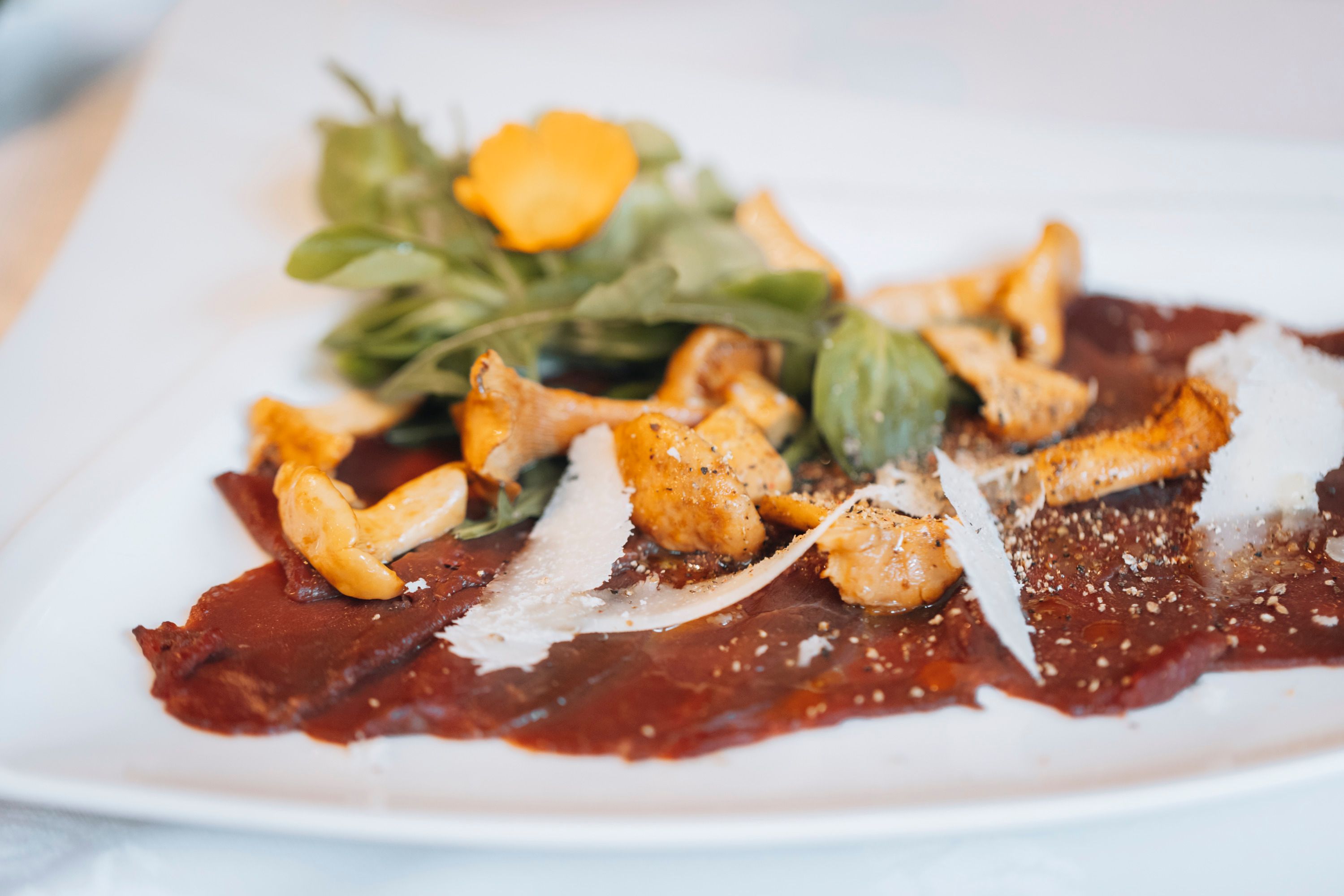 Damhirsch-Carpaccio mit eingelegten Eierschwammerln und Rucola auf einem weißen Teller.