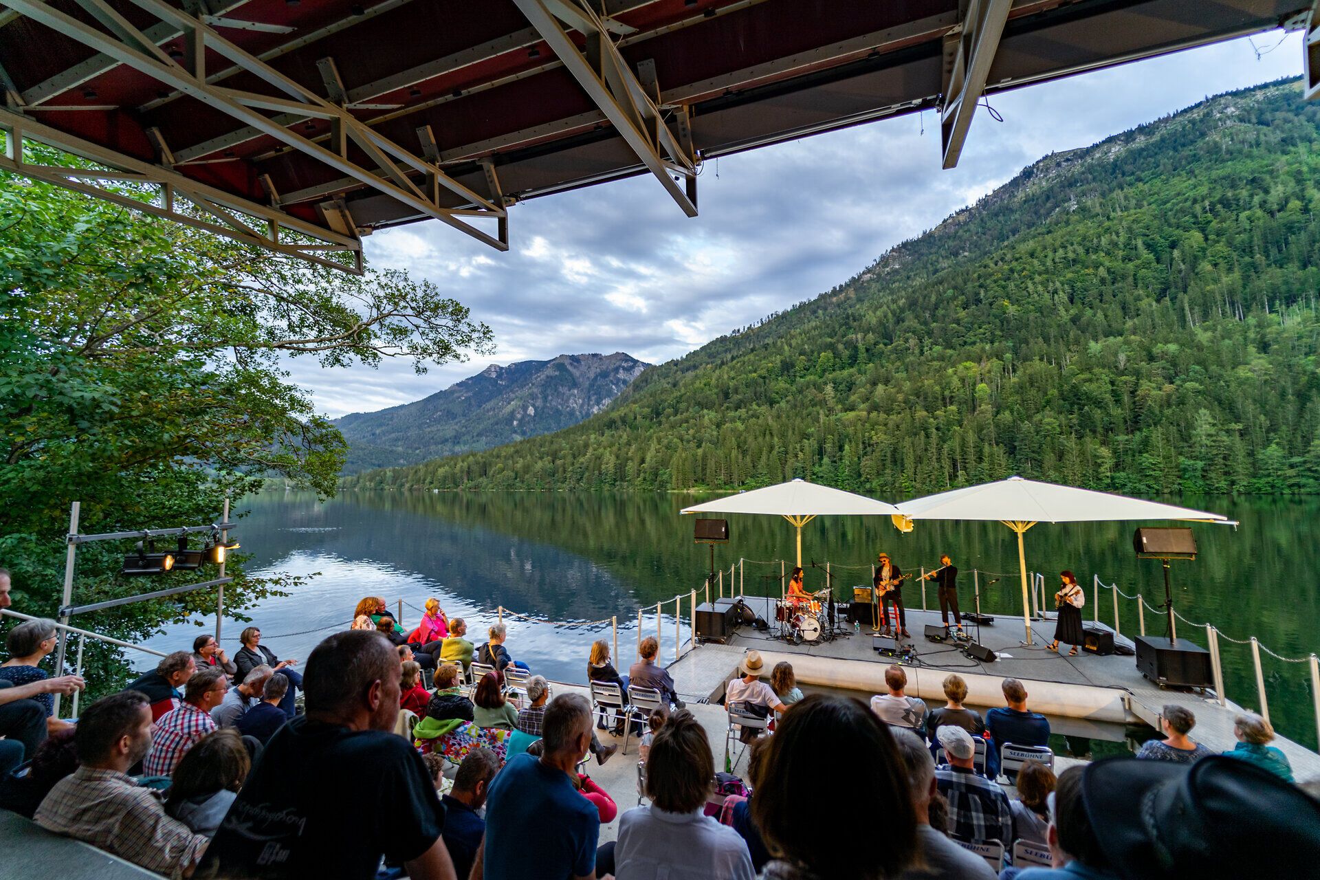 An einem malerischen See, umgeben von majestätischen Bergen, genießen die Besucher ein unvergessliches Konzert unter freiem Himmel. Die sanfte Brise trägt die Klänge der Musik über das Wasser und schafft eine harmonische Verbindung zwischen Natur und Kunst.
