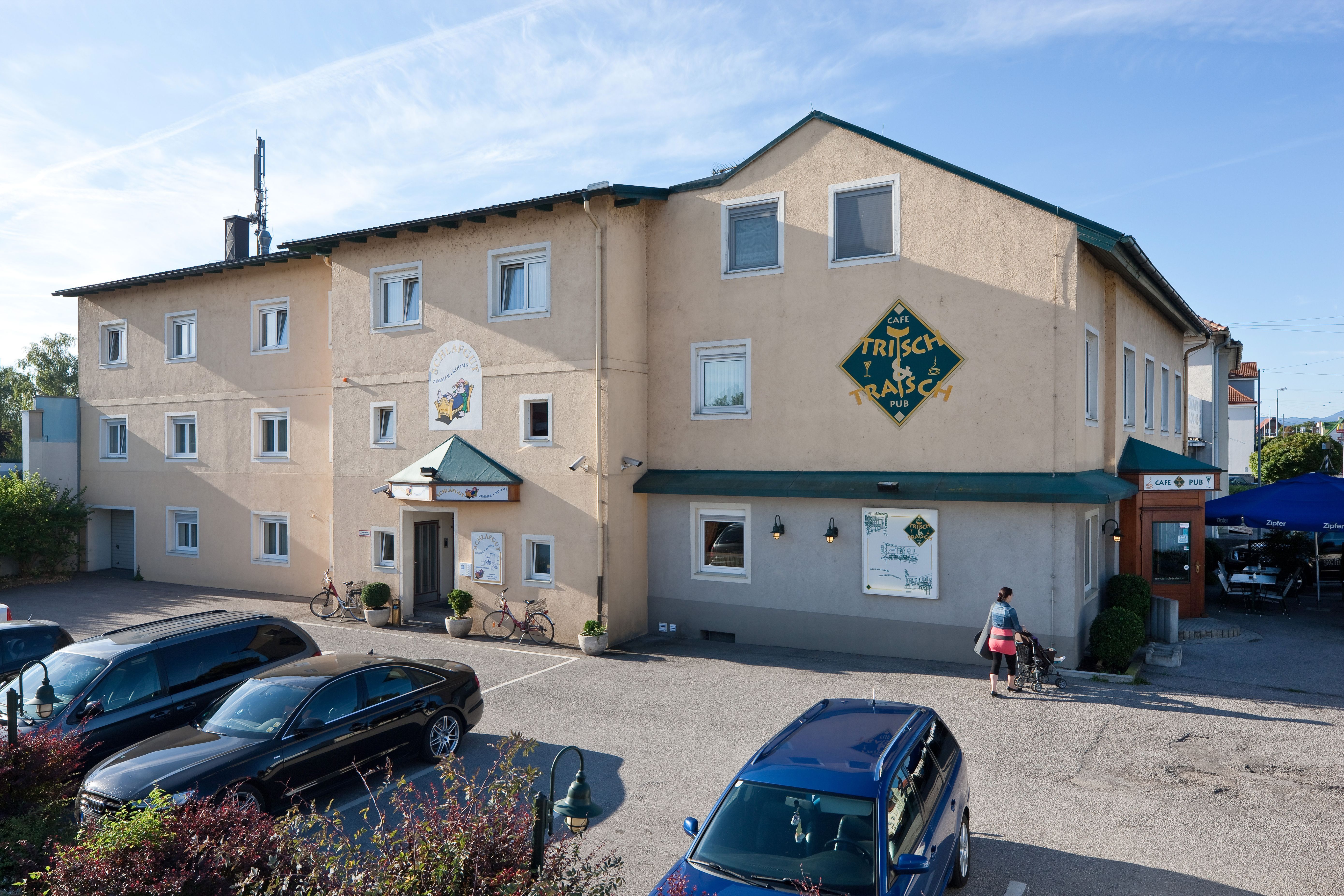 Außenansicht eines Gebäudes mit der Aufschrift 'Pension Schlafgut' und 'Café Trisch & Tratsch'. Autos parken davor, eine Person mit Kinderwagen geht vorbei.