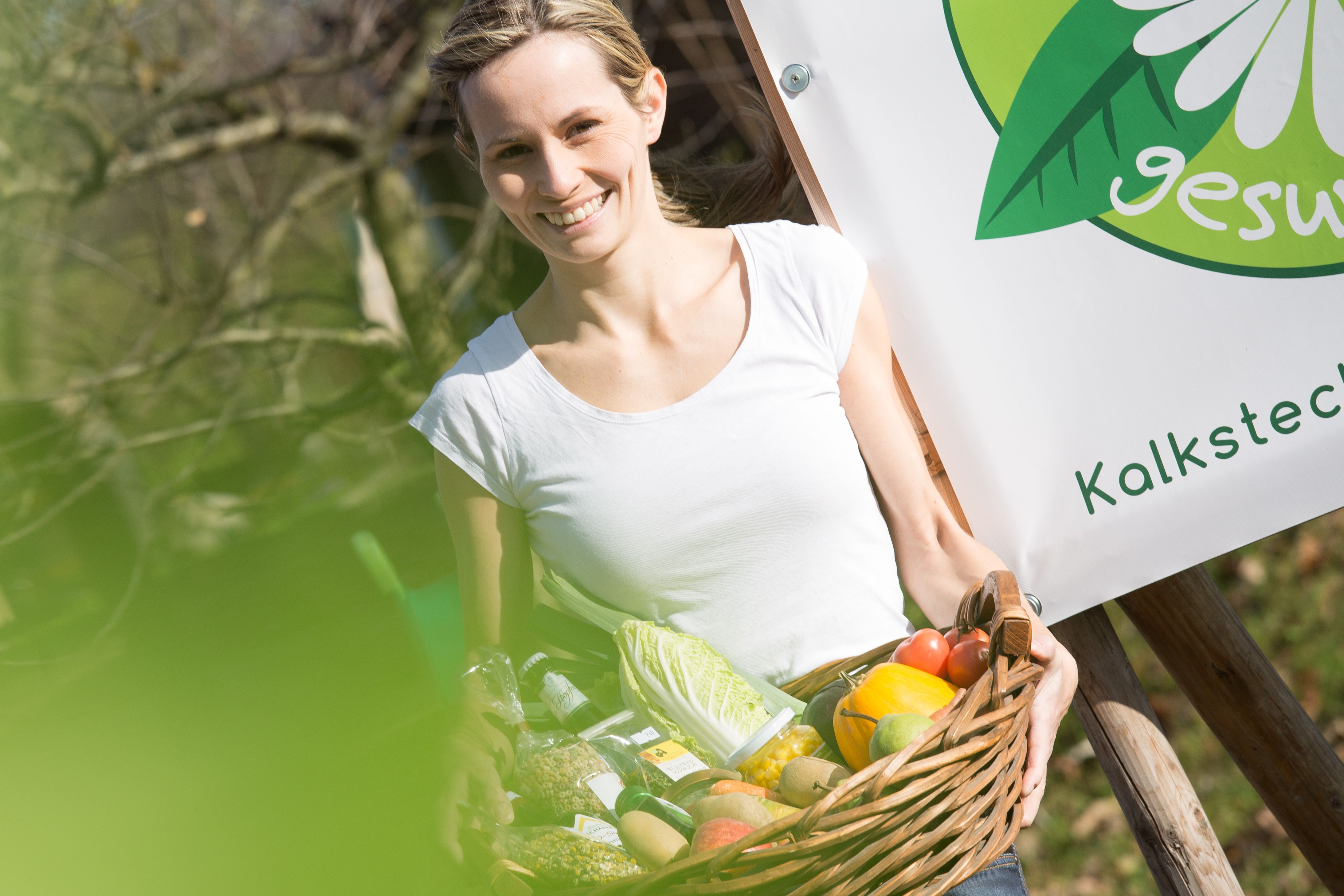 Frau mit Korb voller Gemüse vor einem Schild mit der Aufschrift 'gesund'.