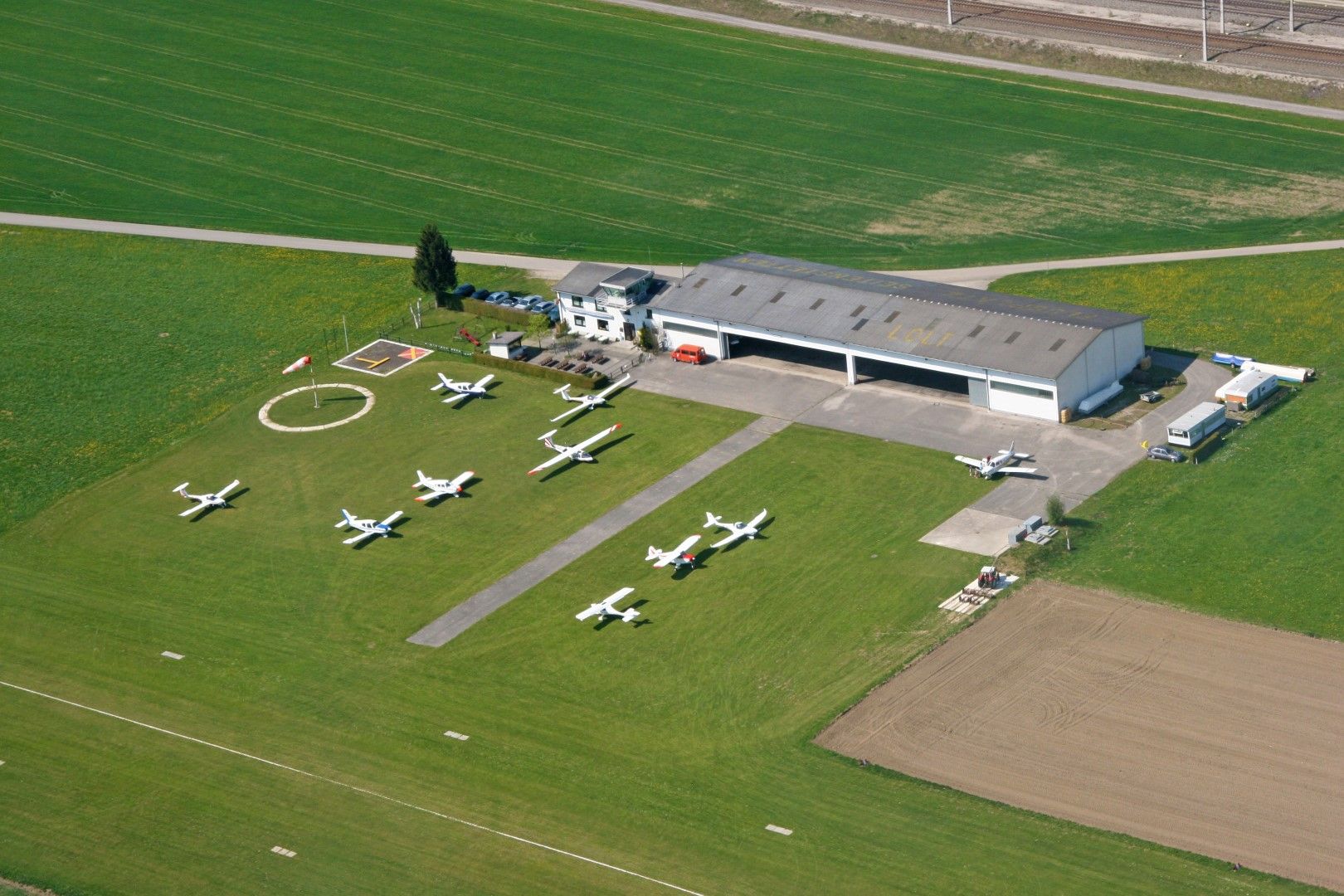 Luftaufnahme eines kleinen Flugplatzes mit mehreren geparkten Flugzeugen und einem Hangar.