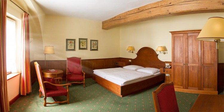 Gemütliches Hotelzimmer mit Holzmöbeln, Doppelbett, zwei roten Sesseln und einem Holztisch. Holzbalken an der Decke, grüne Teppiche und Wandbilder.