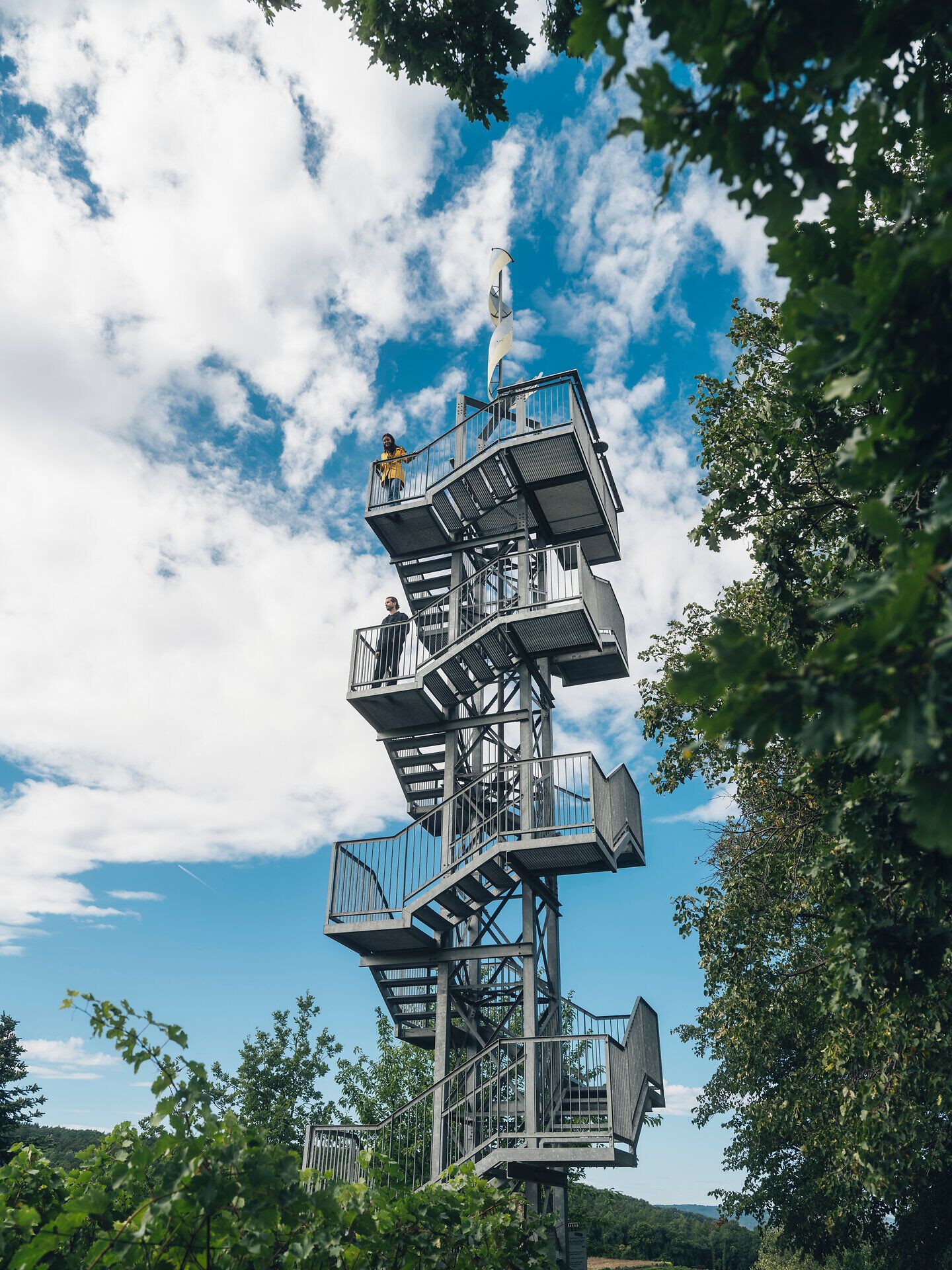 Ein Aussichtsturm ragt zwischen Bäumen und Weinreben in den Himmel, mehrere Personen stehen auf den Plattformen. Die Aufnahme entstand beim Weingut und der Vinothek Dam in der Ahrenberger und Eichberger Kellergasse im Traisental, Niederösterreich.