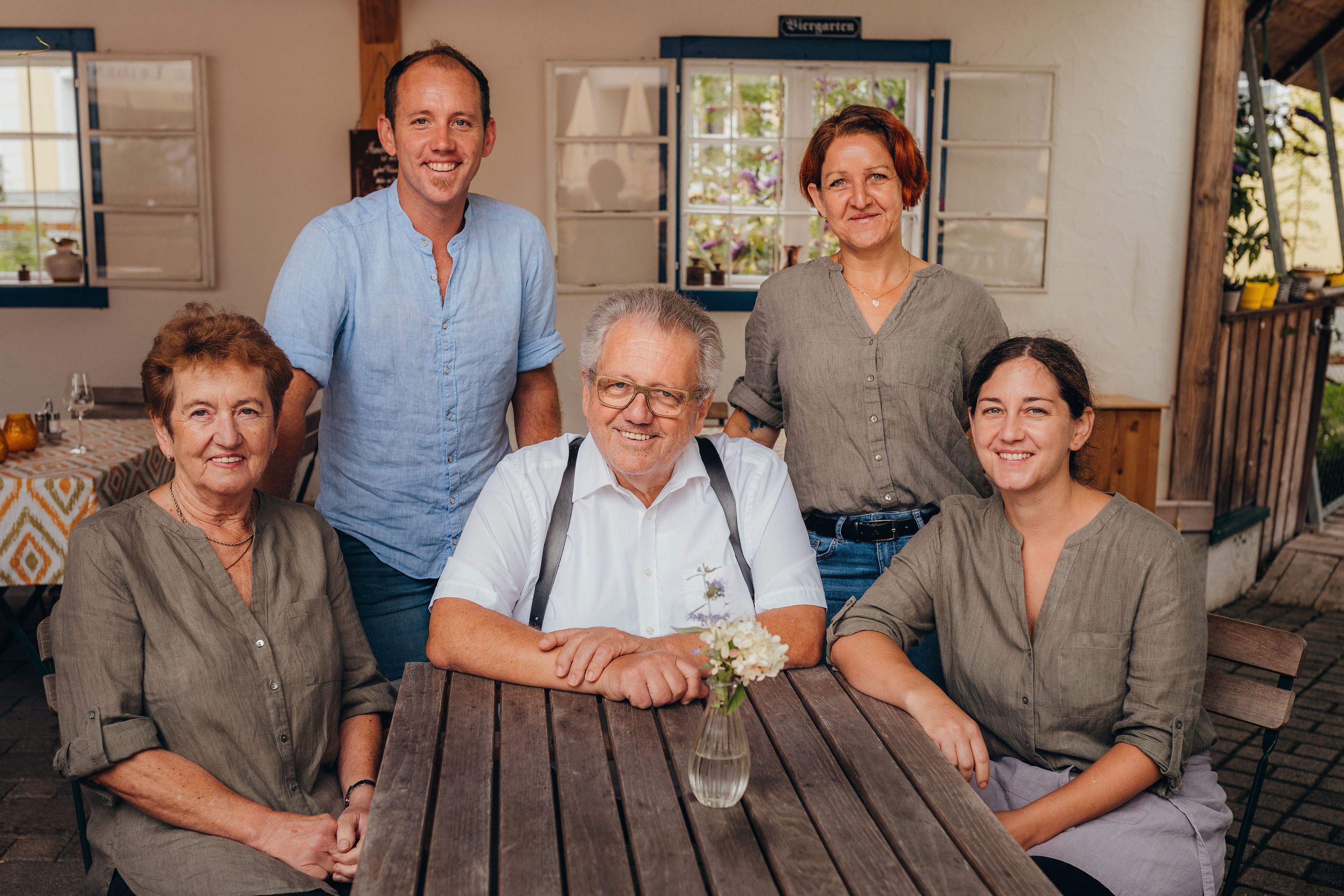 Fünf Personen sitzen und stehen um einen Holztisch in einem überdachten Außenbereich.