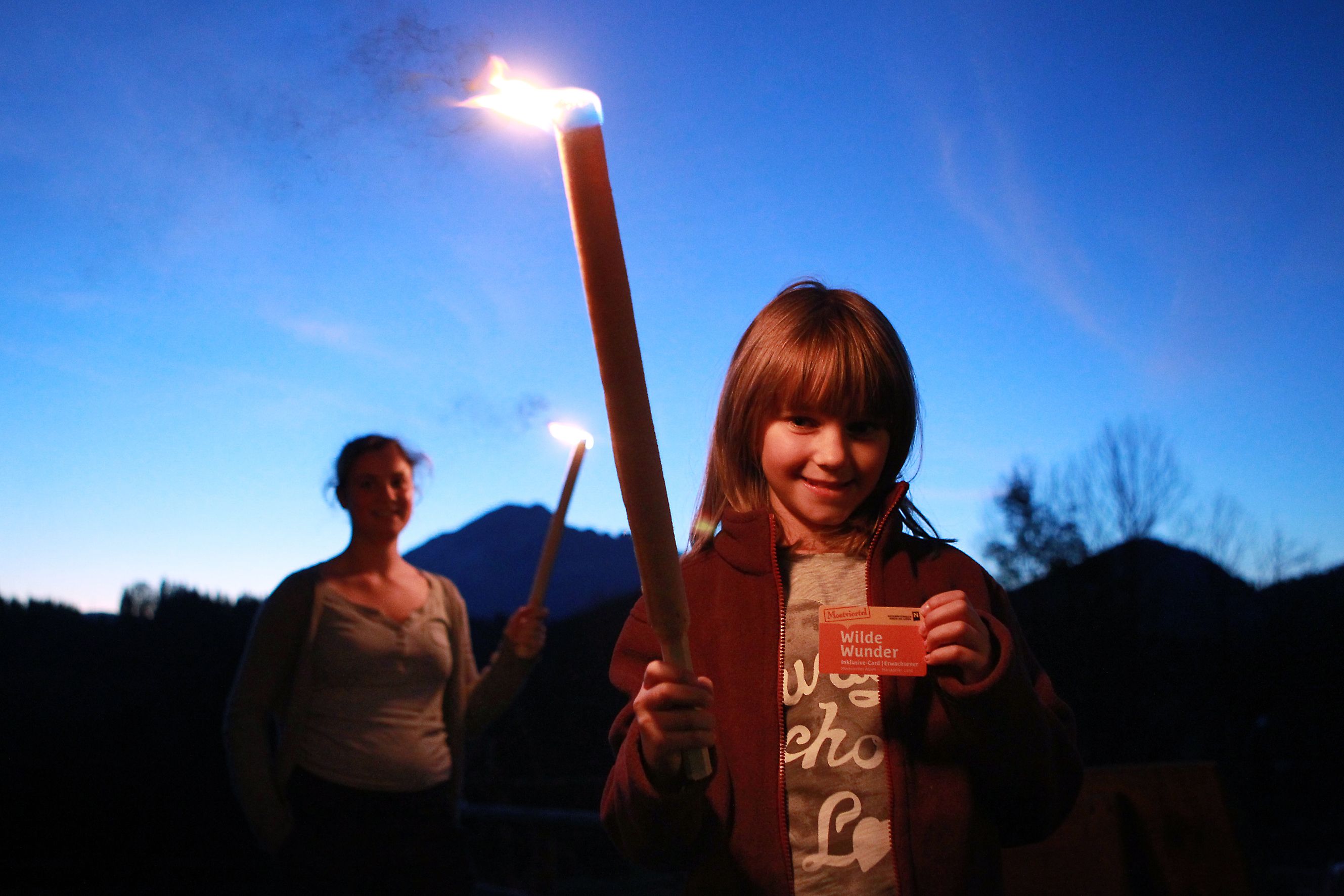 Ein Kind hält eine brennende Fackel und eine Karte, im Hintergrund eine Frau mit Fackel, vor einer Bergkulisse bei Dämmerung.