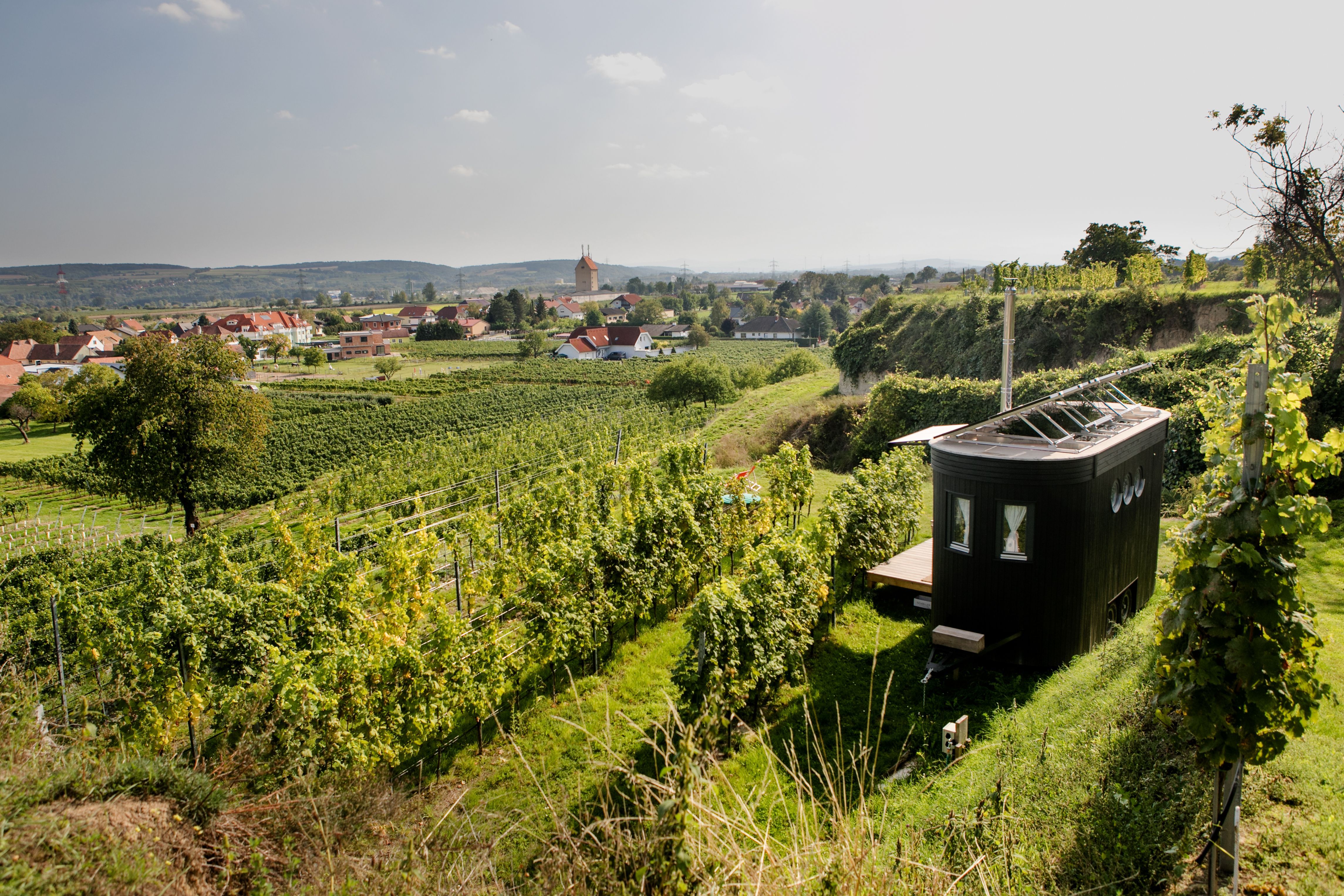 Ein kleiner, moderner Wohnwagen steht inmitten von Weinbergen mit Blick auf ein Dorf und Hügel im Hintergrund.