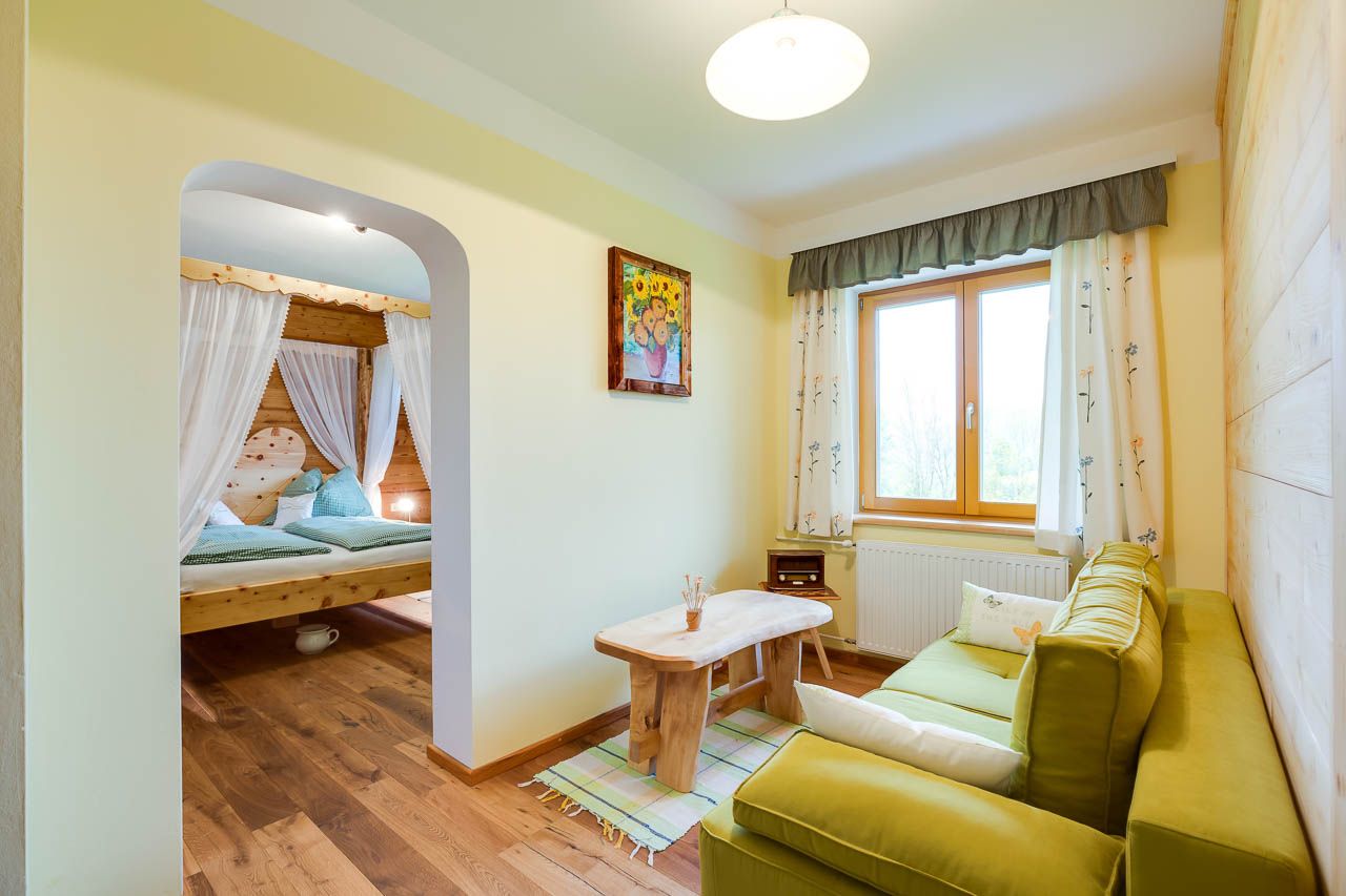 Gemütliches Zimmer mit Holzmöbeln, grünem Sofa, großem Fenster und einem Himmelbett im Hintergrund.