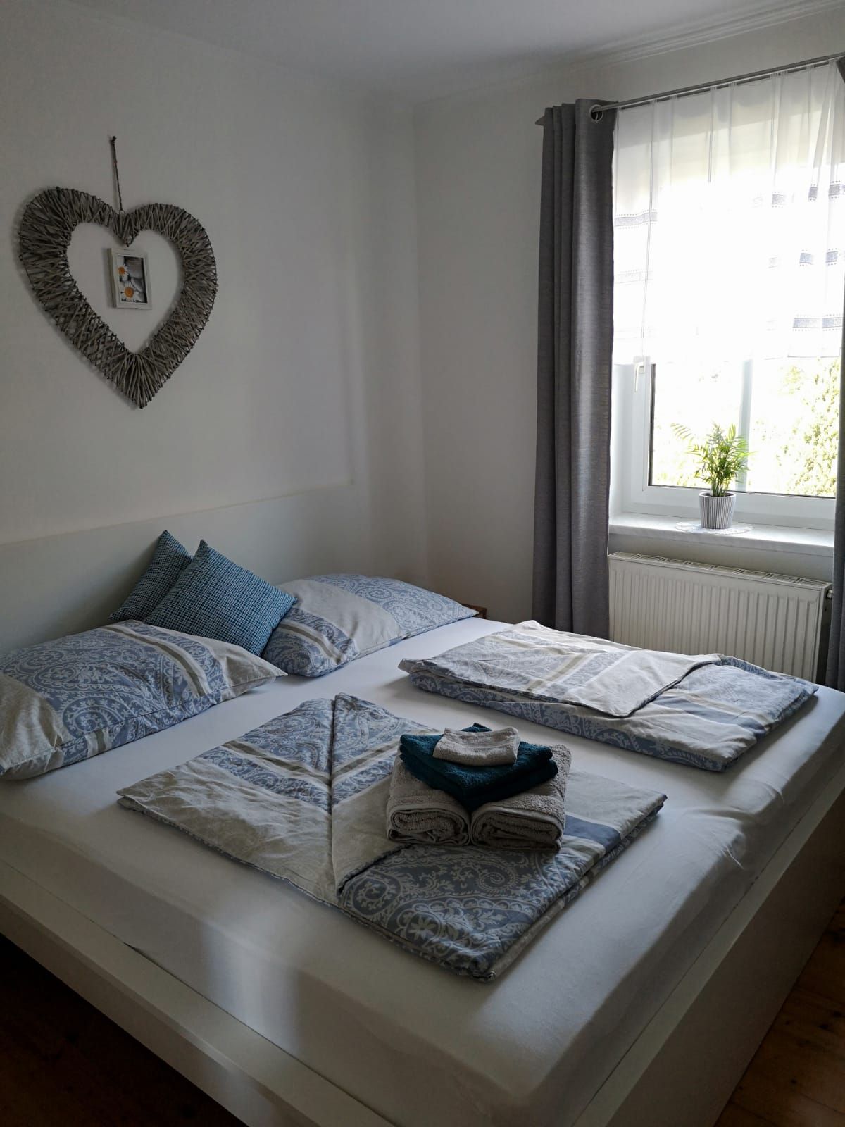 Ein gemütliches Schlafzimmer mit einem Doppelbett, dekoriert mit blauer Bettwäsche und Handtüchern. Ein Herzbild hängt an der Wand, ein Fenster mit Vorhang daneben.