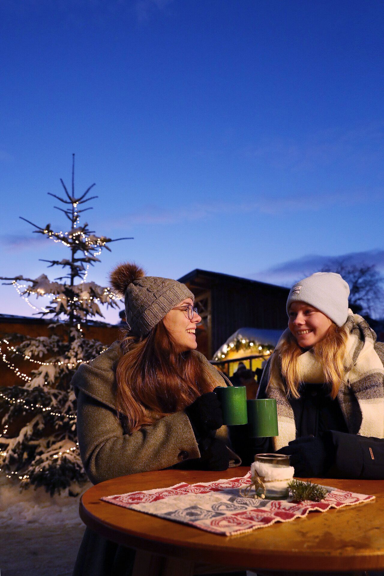 Zwei fröhliche Frauen genießen heiße Getränke in der winterlichen Atmosphäre eines Adventmarktes. Umgeben von festlich geschmückten Bäumen und einem klaren, blauen Himmel, strahlt die Szene Wärme und Gemütlichkeit aus. Der Duft von Glühwein und frisch gebackenem Gebäck liegt in der Luft und lädt zum Verweilen ein.