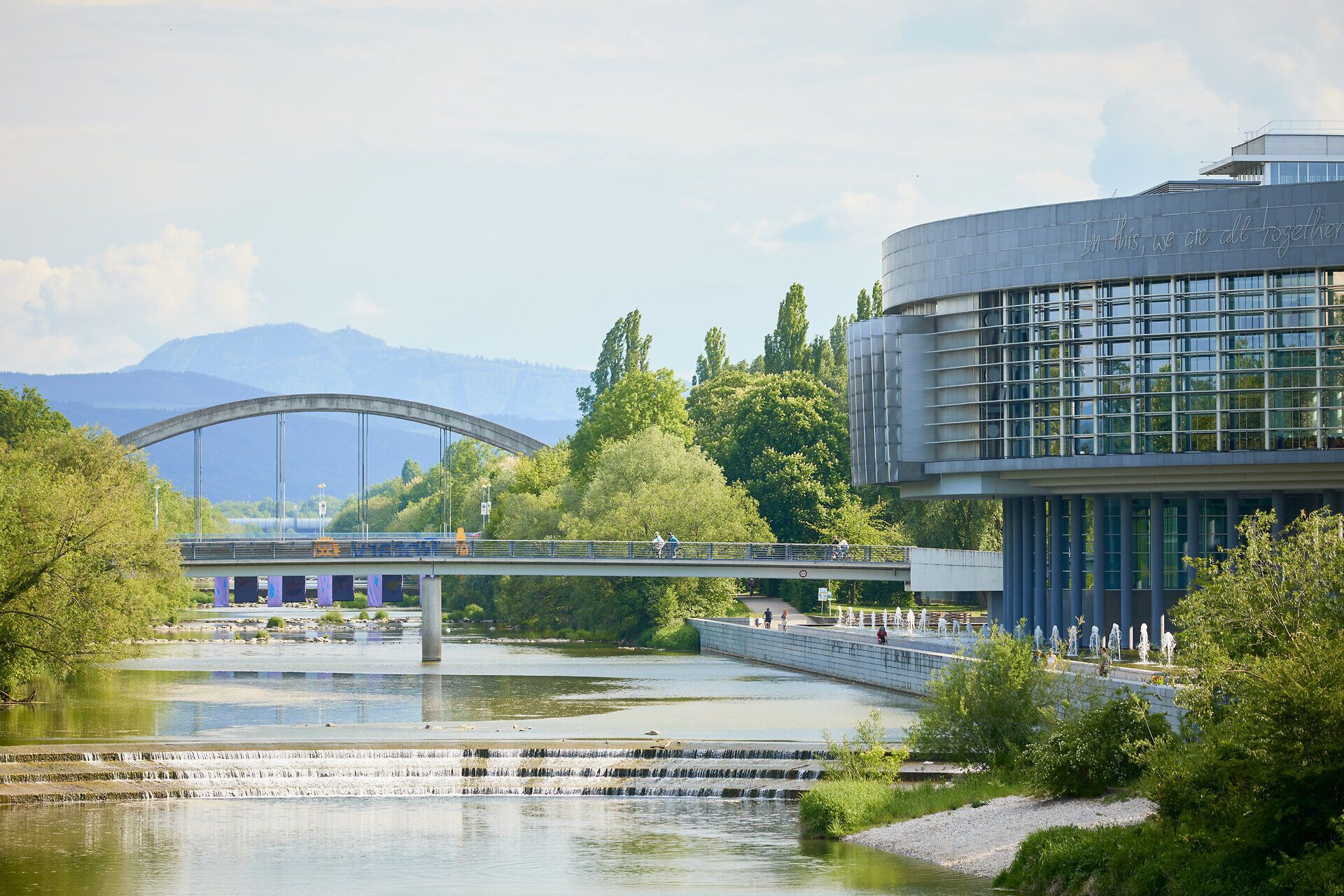 Die sanften Wellen des Wassers spiegeln die grüne Pracht der Bäume wider, während die Brücke elegant über den Fluss führt. Hier, wo Natur und Architektur harmonisch verschmelzen, lädt die Umgebung zu einem entspannten Spaziergang ein. Genießen Sie die frische Luft und die malerische Aussicht auf die Stadt.