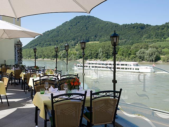 Terrasse mit gedeckten Tischen, Blick auf Fluss und vorbeifahrendes Schiff, umgeben von grünen Hügeln.