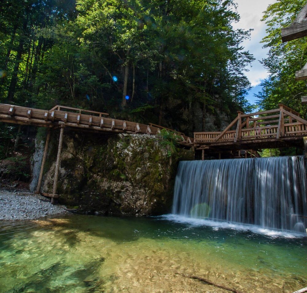 Ein sanfter Wasserfall plätschert in das klare, grüne Wasser, umgeben von üppigem Wald. Die Holzbrücke lädt dazu ein, die malerische Umgebung zu erkunden und die frische Bergluft zu genießen. Hier, in der Erlebniswelt Mendlingtal, wird jeder Schritt zum Abenteuer für die ganze Familie.