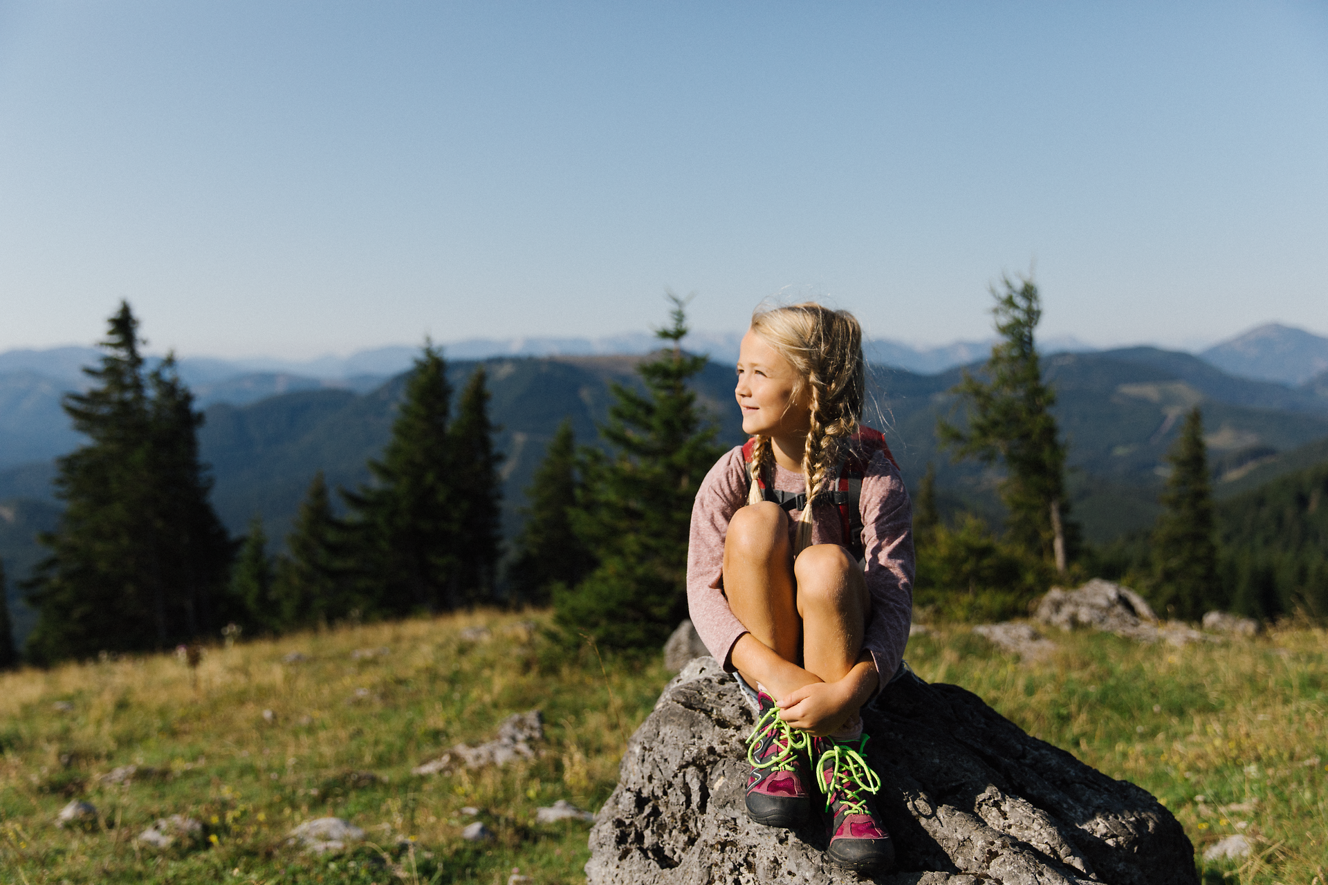Ein strahlendes Kind sitzt auf einem großen Stein und genießt die atemberaubende Aussicht auf die umliegenden Berge. Die sanften Hügel und der klare Himmel schaffen eine perfekte Kulisse für unvergessliche Familienabenteuer in der Natur.