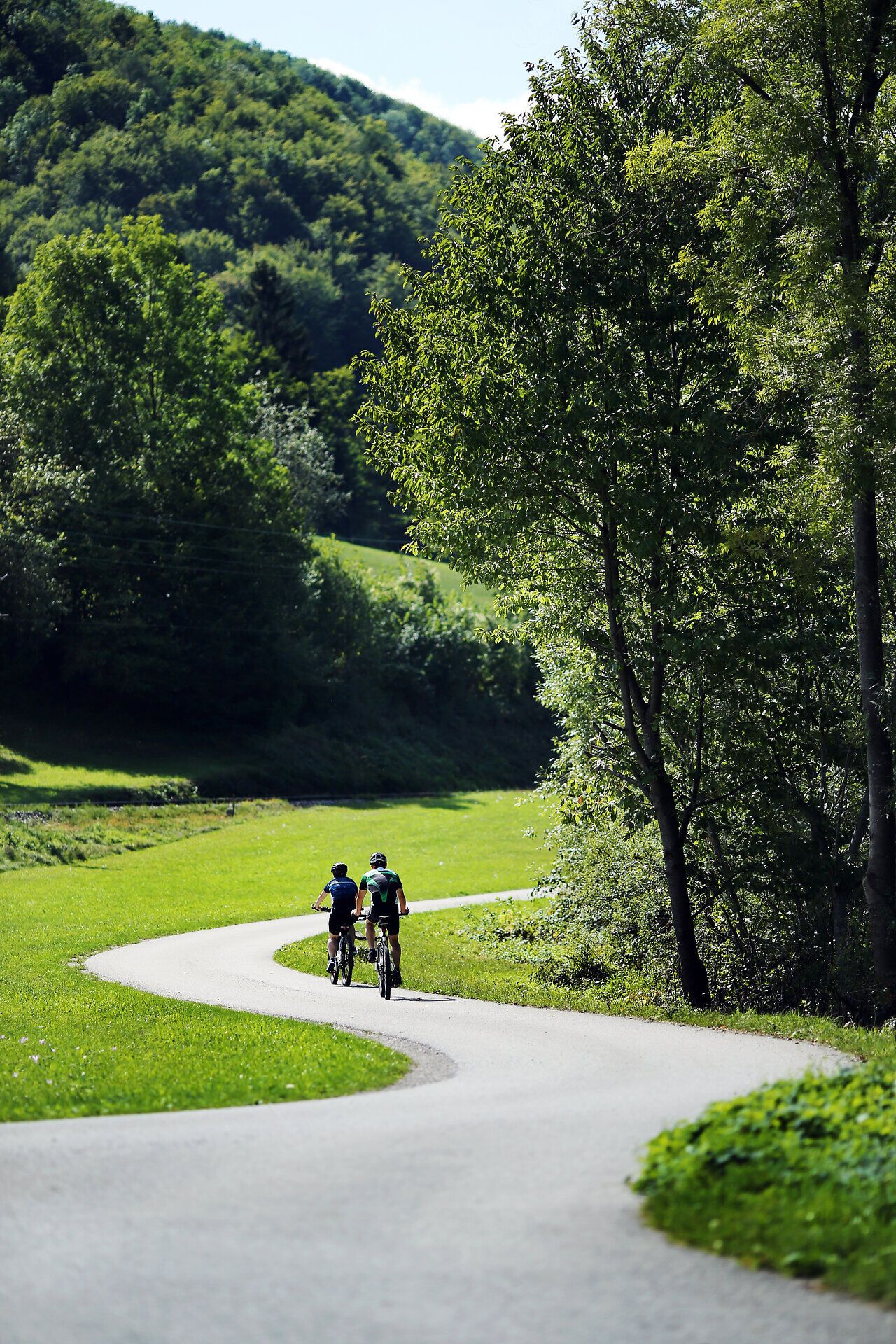 Sanfte Hügel umgeben den malerischen Radweg, der durch eine grüne Landschaft führt. Zwei Radfahrer genießen die frische Luft und die Ruhe der Natur, während sie die Schönheit der Umgebung in vollen Zügen erleben.