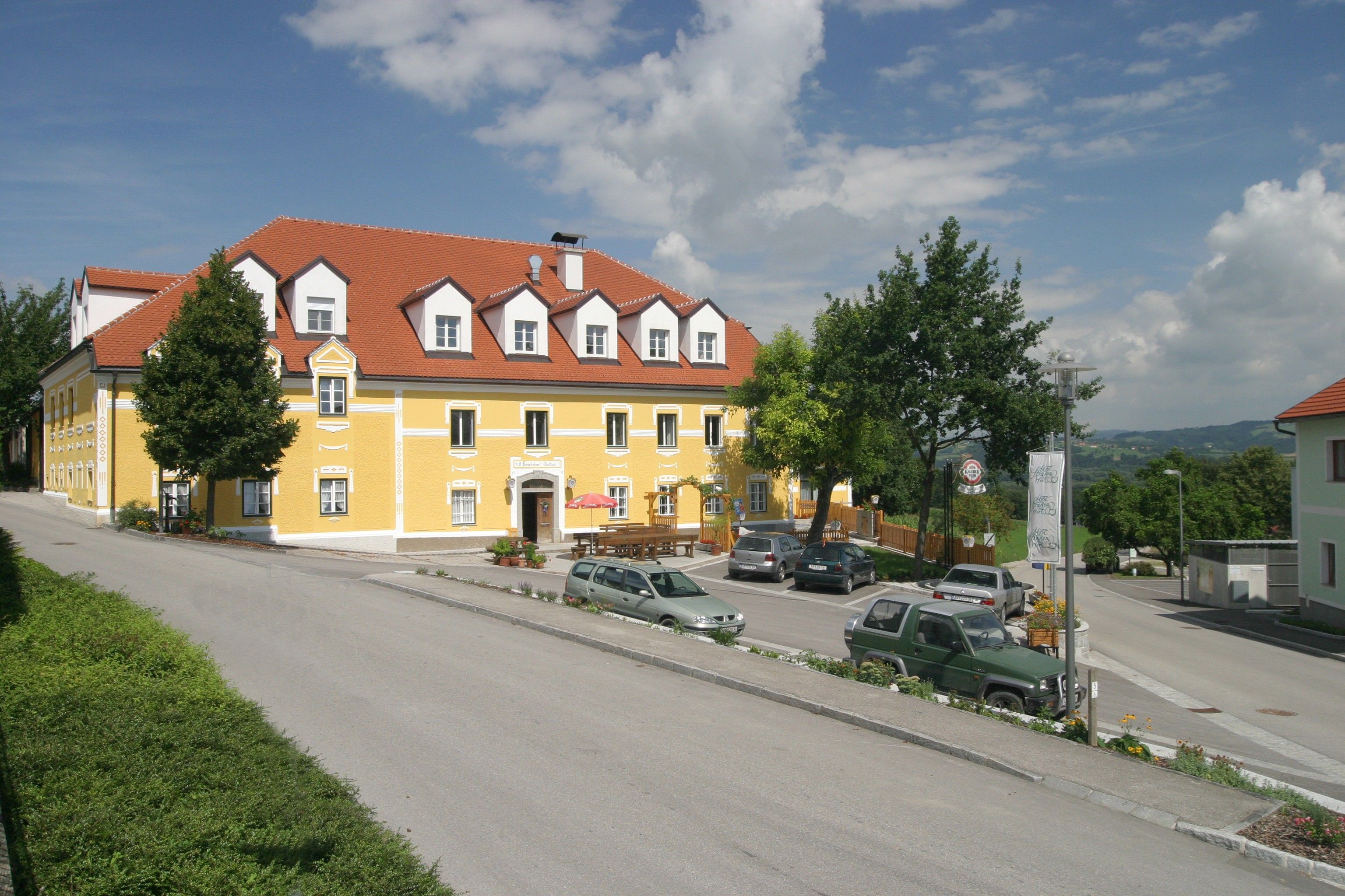 Ein gelbes Gebäude mit rotem Dach, umgeben von Autos und Bäumen, unter blauem Himmel.