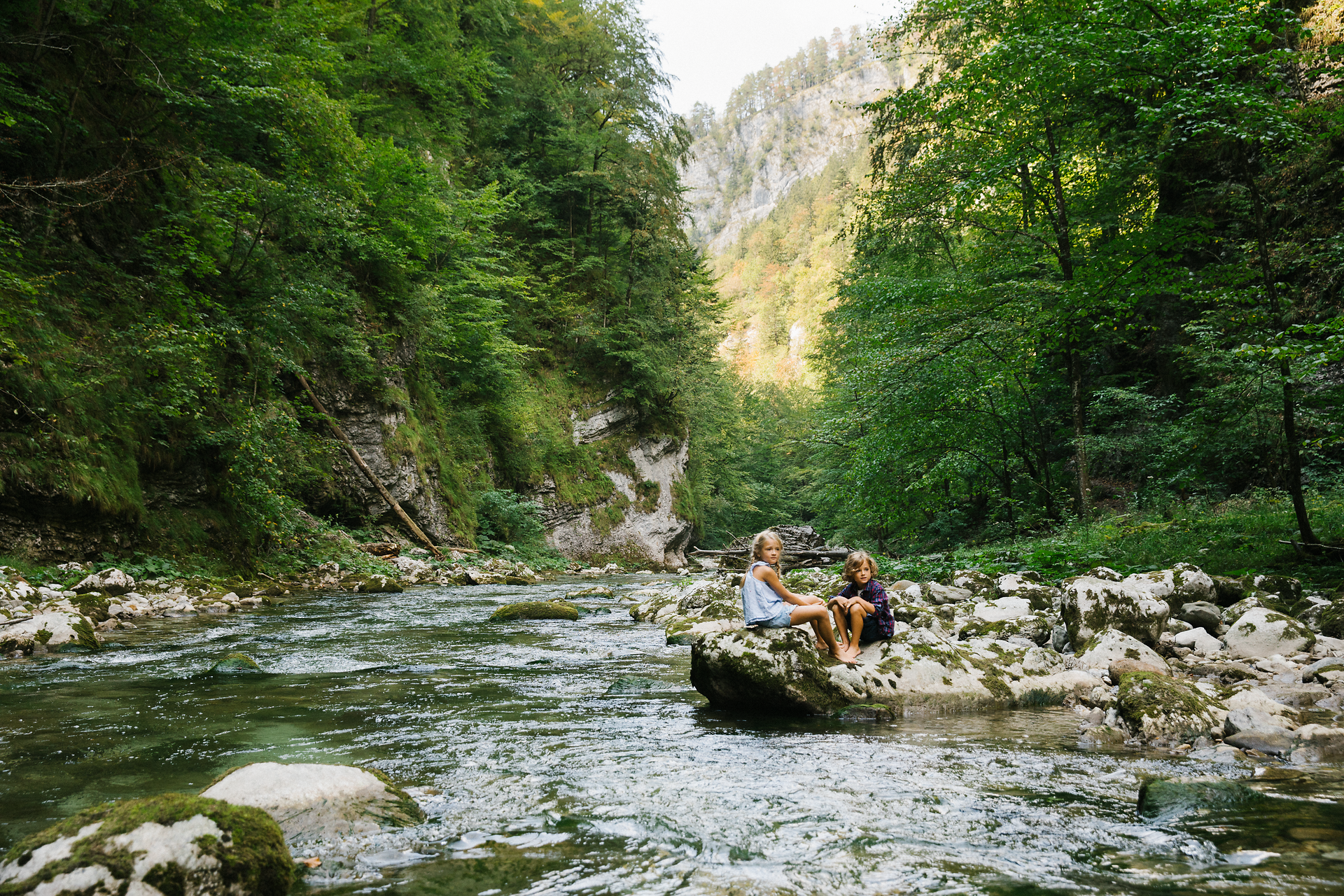 Ein erfrischender Fluss schlängelt sich durch eine grüne Landschaft, umgeben von hohen Bäumen und sanften Hügeln. Familien genießen die Ruhe der Natur, während sie auf den glatten Steinen am Ufer sitzen und die kühle Brise spüren. Hier wird der Sommer in den Bergen zu einem unvergesslichen Erlebnis.