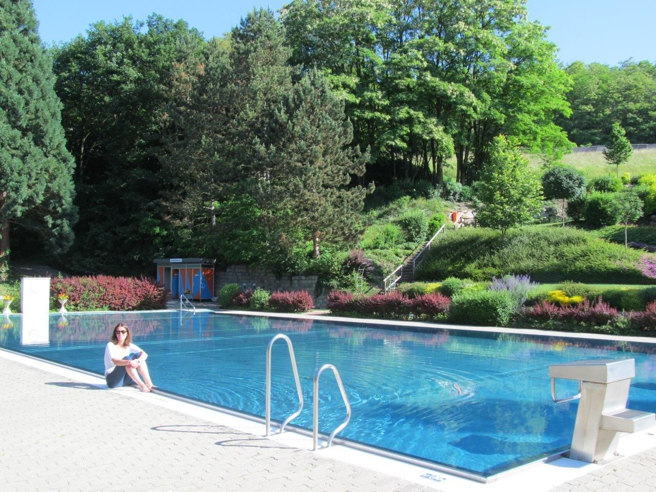 Frau sitzt am Rand eines Freibads mit Wald im Hintergrund.