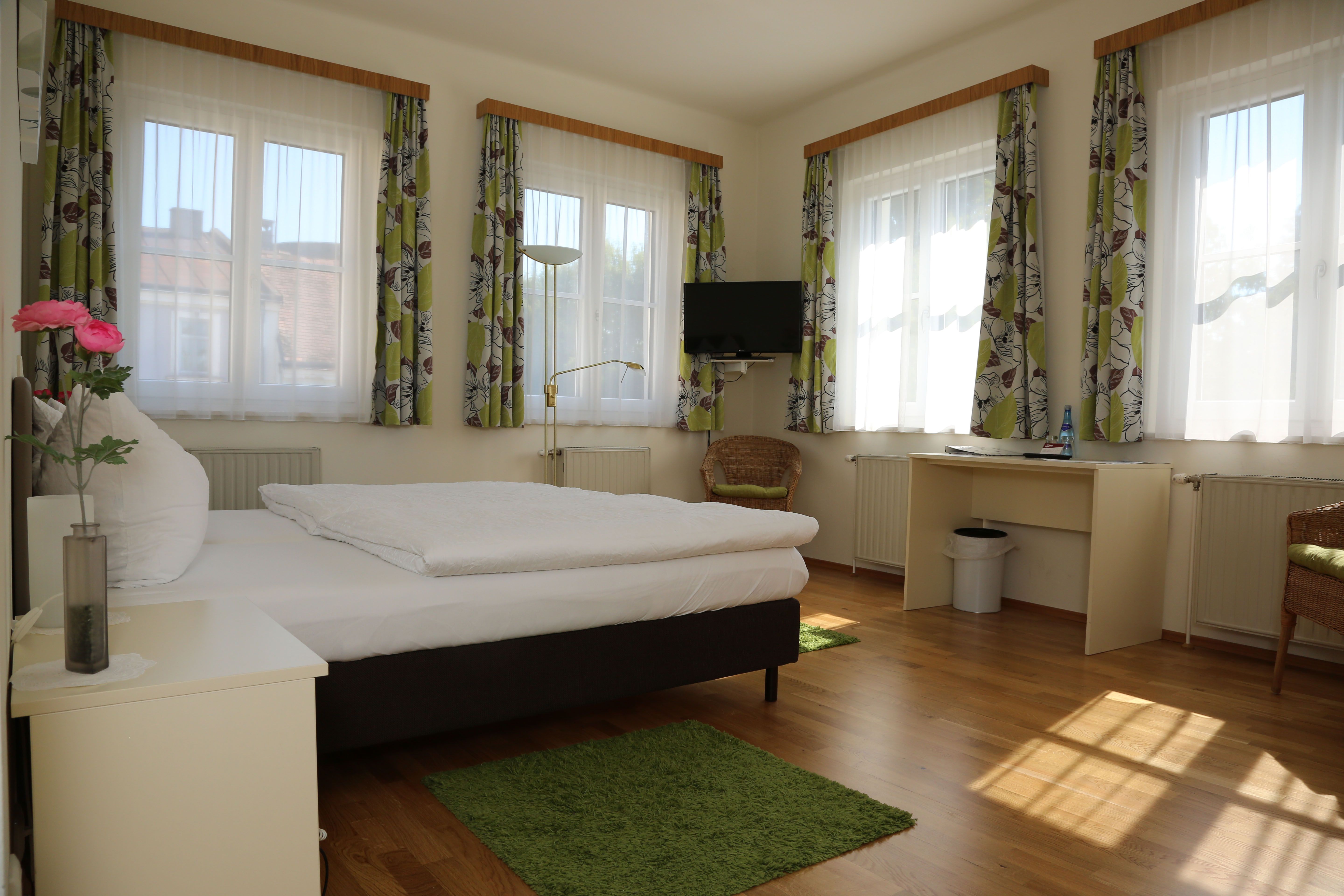 Helles Zimmer mit Bett, Schreibtisch, Fernseher und mehreren Fenstern mit Vorhängen.