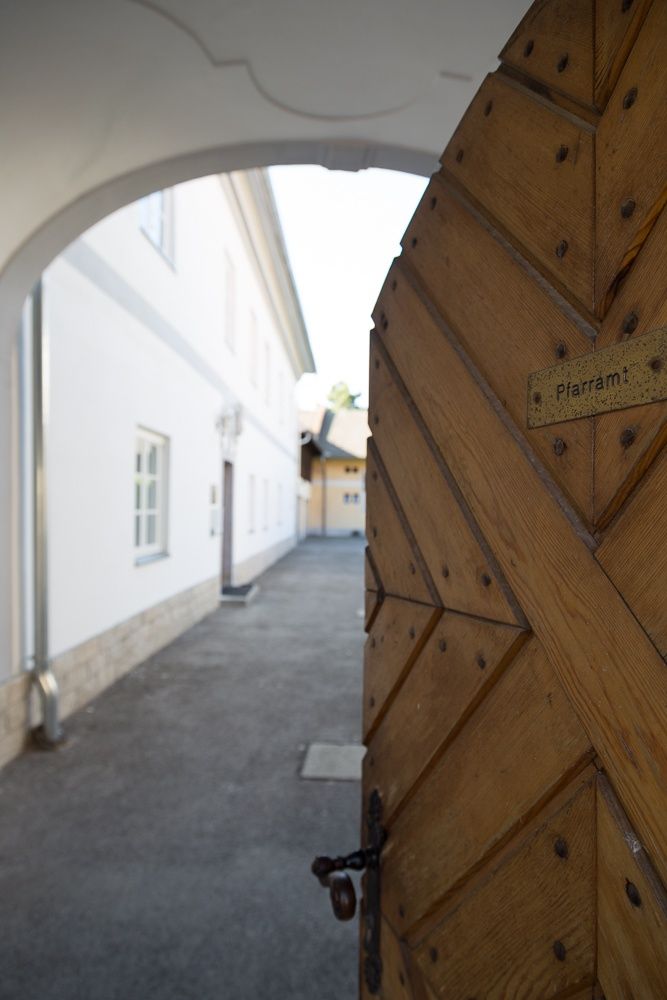 Ein hölzernes Tor mit der Aufschrift 'Pfarramt' öffnet sich zu einem Innenhof mit weißen Gebäuden.