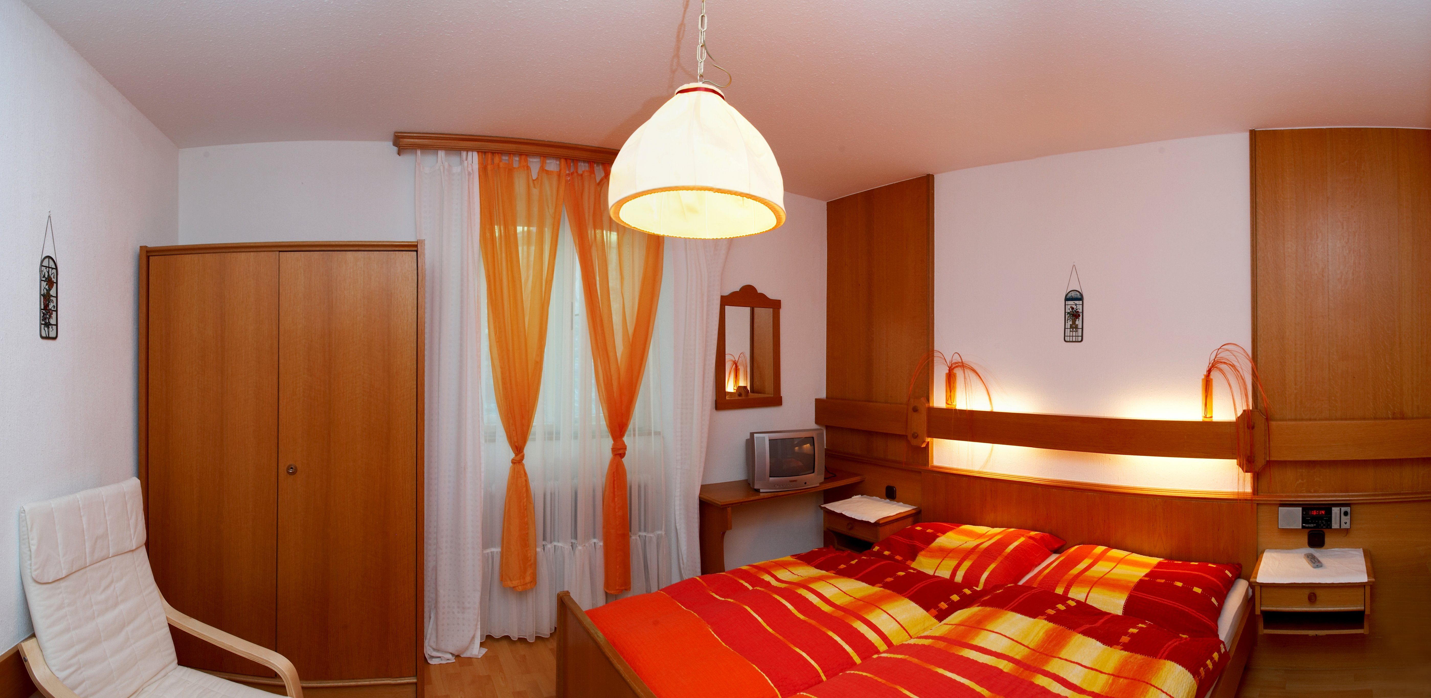 Gemütliches Hotelzimmer mit Doppelbett, Holzschrank, Fernseher und orange-weißen Vorhängen.