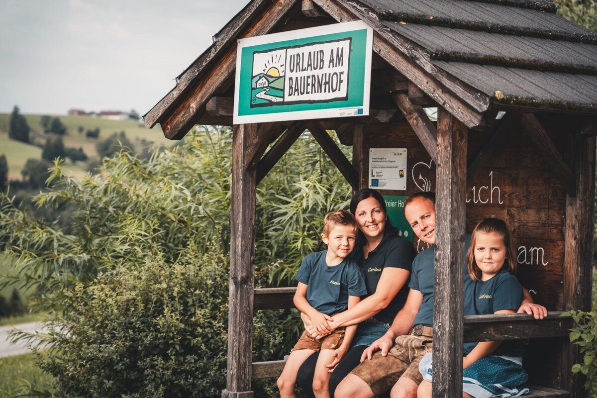 Familie Korntheuer sitzt in einem Holzunterstand mit einem Schild 'Urlaub am Bauernhof'. Im Hintergrund sind grüne Hügel zu sehen.