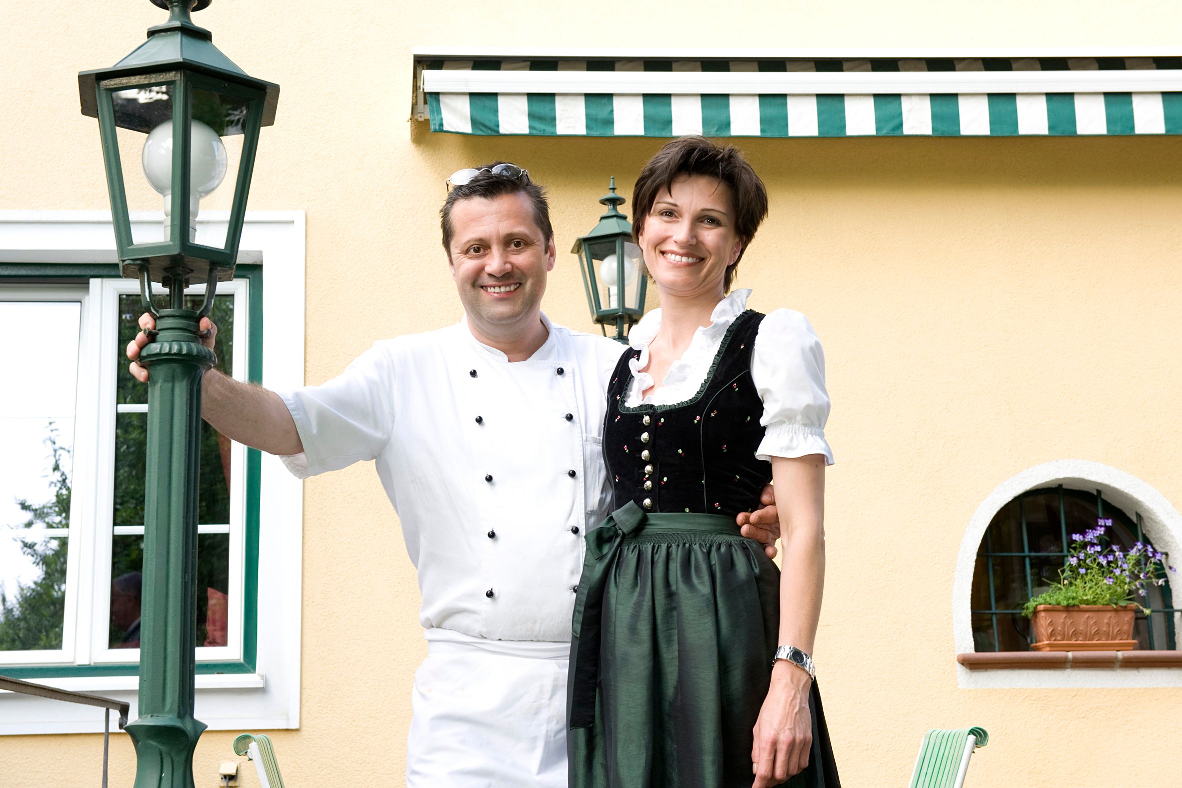 Ein Mann in Kochkleidung und eine Frau in traditioneller Tracht stehen lächelnd vor einem gelben Gebäude mit grünen Laternen.