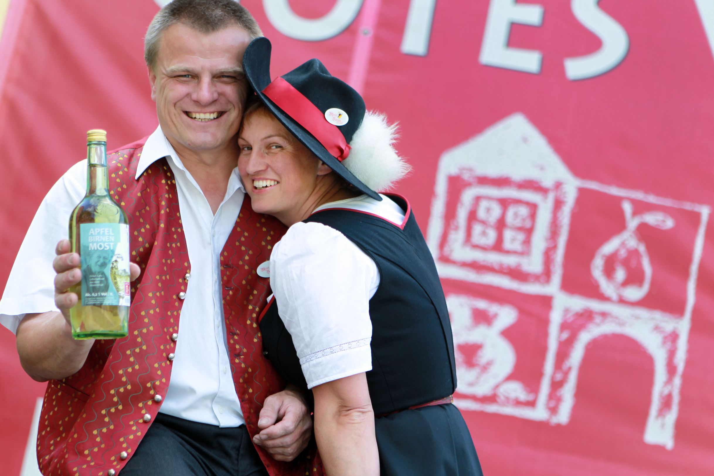 Ein Mann und eine Frau in traditioneller Kleidung lächeln und halten eine Flasche Apfel-Birnen-Most vor einem roten Hintergrund.