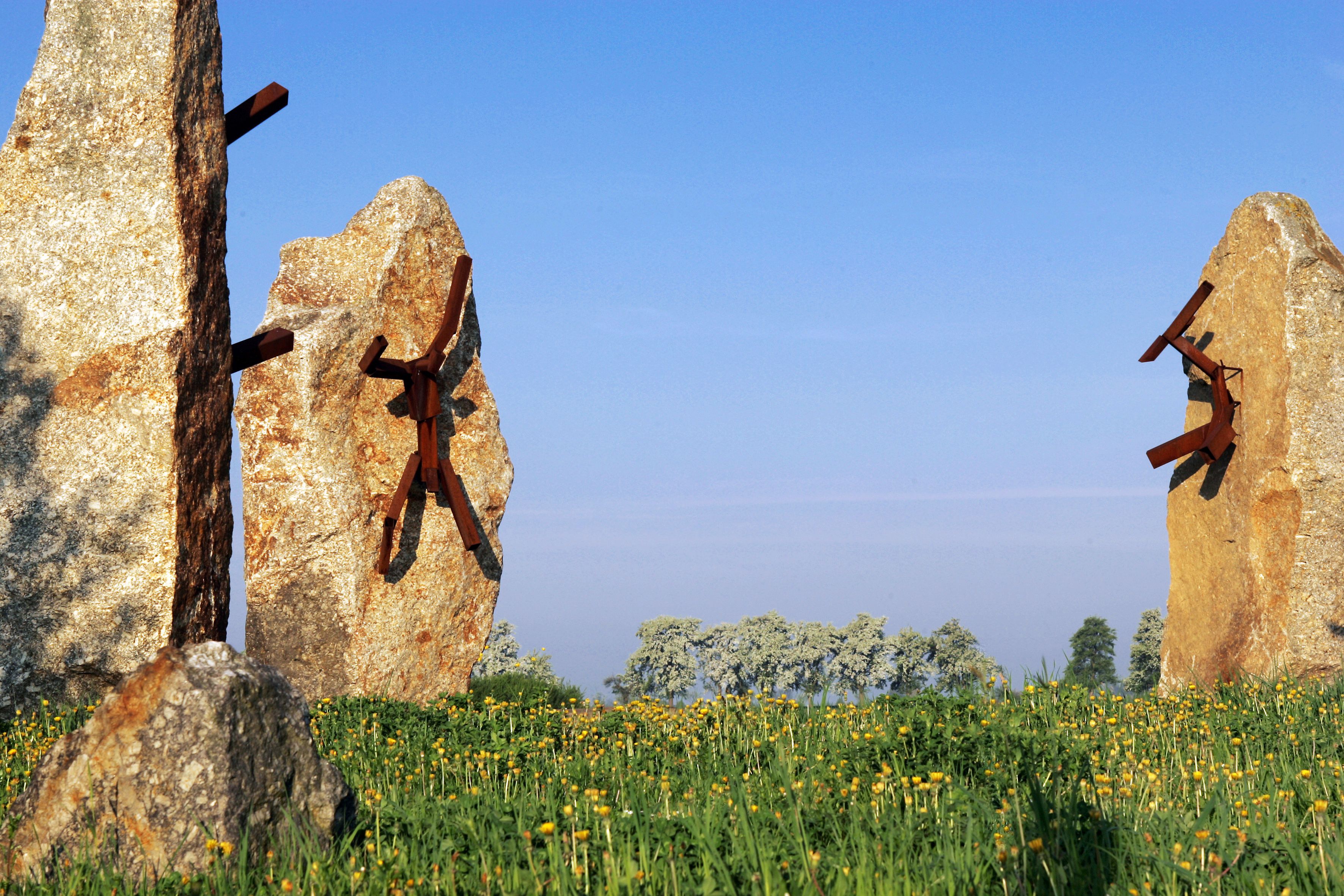 Steinskulpturen mit Metallfiguren auf einer Wiese unter blauem Himmel.