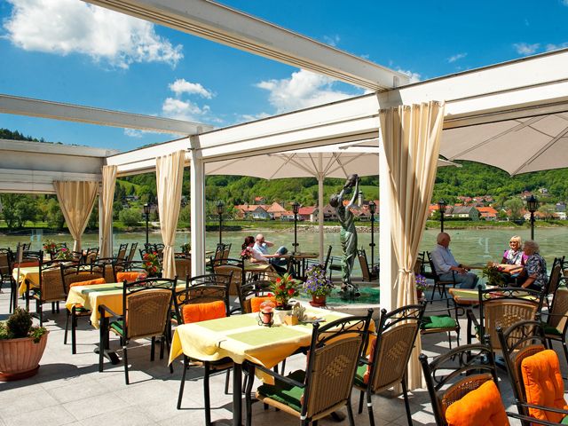 Restaurantterrasse mit Blick auf einen Fluss und grüne Hügel im Hintergrund.