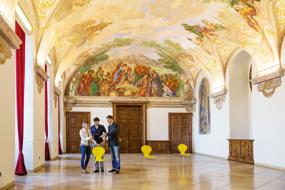 Innenansicht eines prunkvollen Raumes im Stift Göttweig mit Fresken an der Decke und drei Personen, die sich unterhalten.