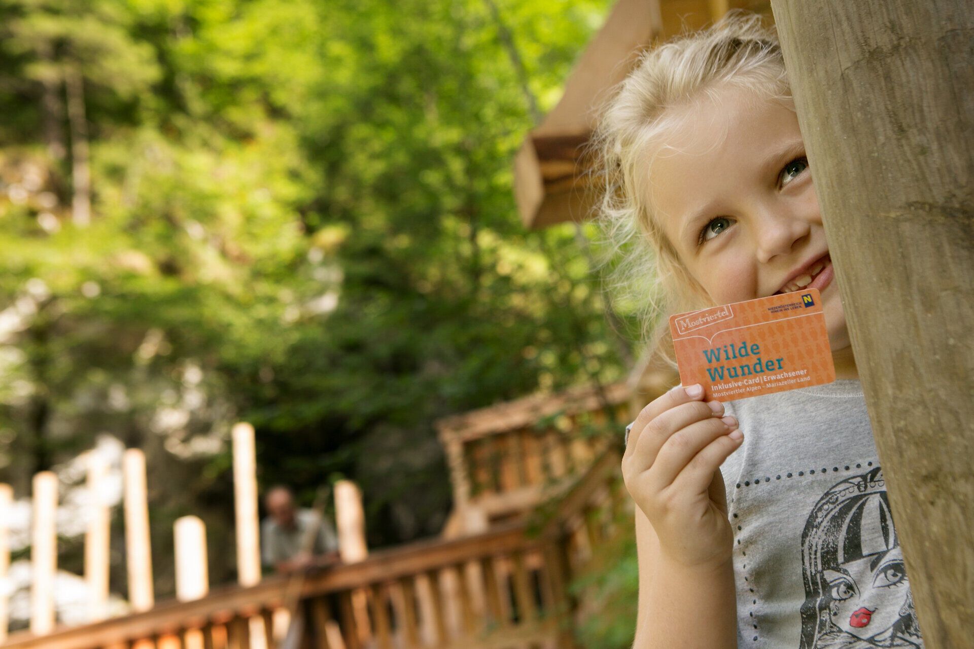 Ein fröhliches Mädchen lächelt, während sie die Wilde Wunder Card in der Hand hält, umgeben von der üppigen Natur des Mendlingtals. Die sanften Hügel und das dichte Grün schaffen eine einladende Atmosphäre für Familien, die unvergessliche Abenteuer in dieser malerischen Landschaft erleben möchten.