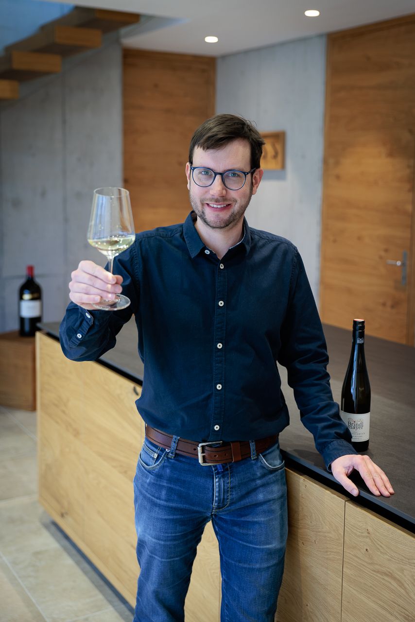 Ein Mann mit Brille hält ein Glas Weißwein in einem modernen Raum mit Holzelementen.