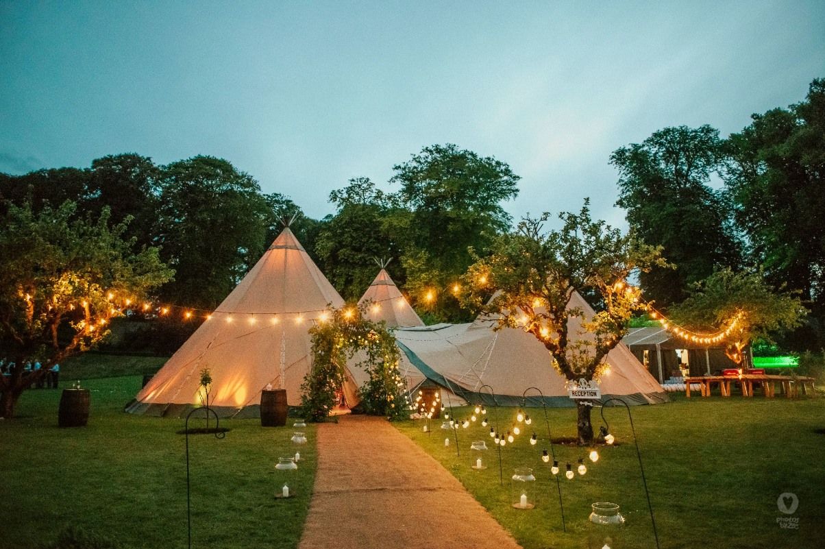 Ein beleuchtetes Tipi-Zelt in einem Garten mit Lichterketten und Bäumen bei Dämmerung.