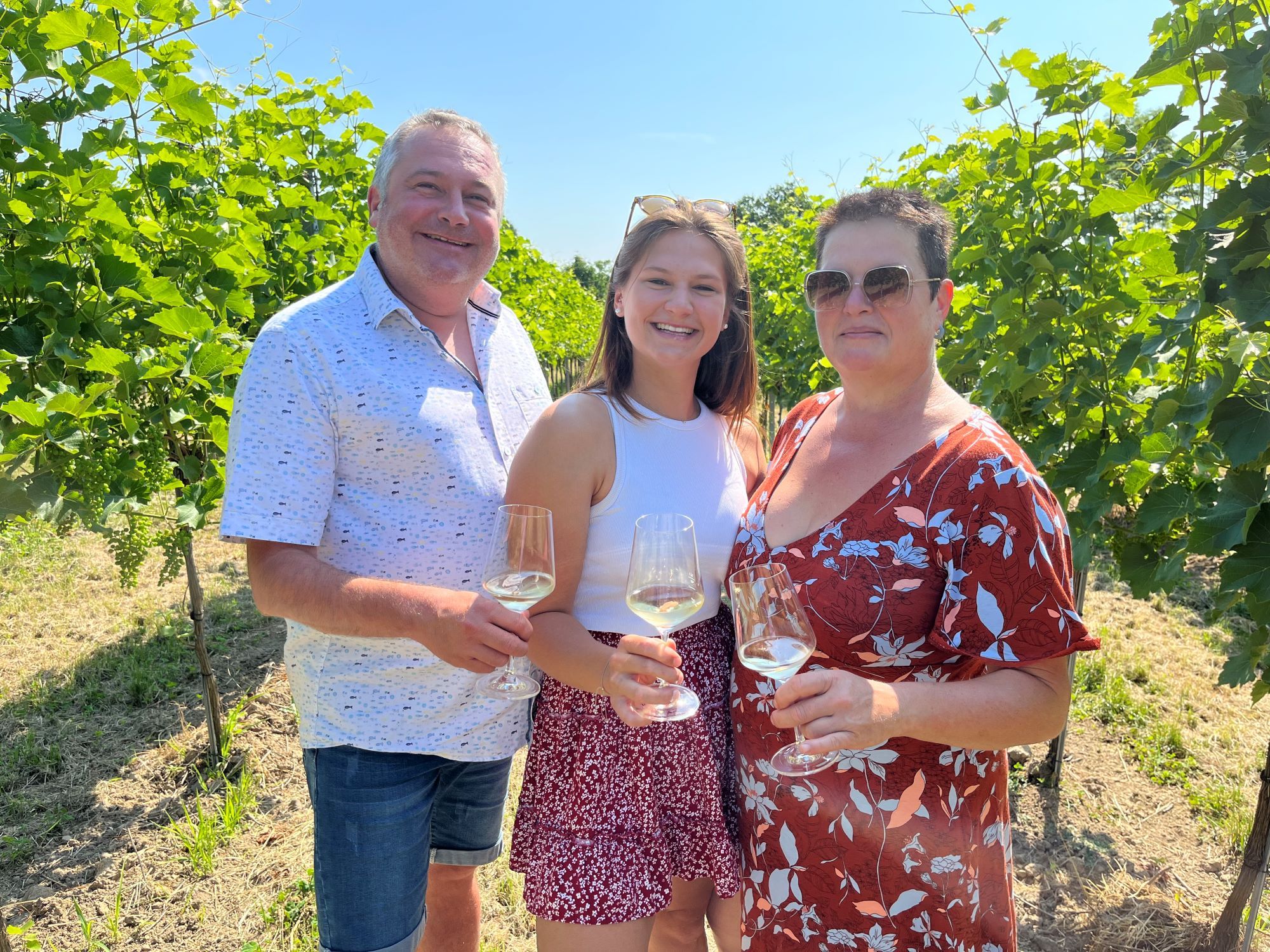 Familie Heimel mit Weingläsern in der Hand. Die Familie steht zwischen Weinreben. 