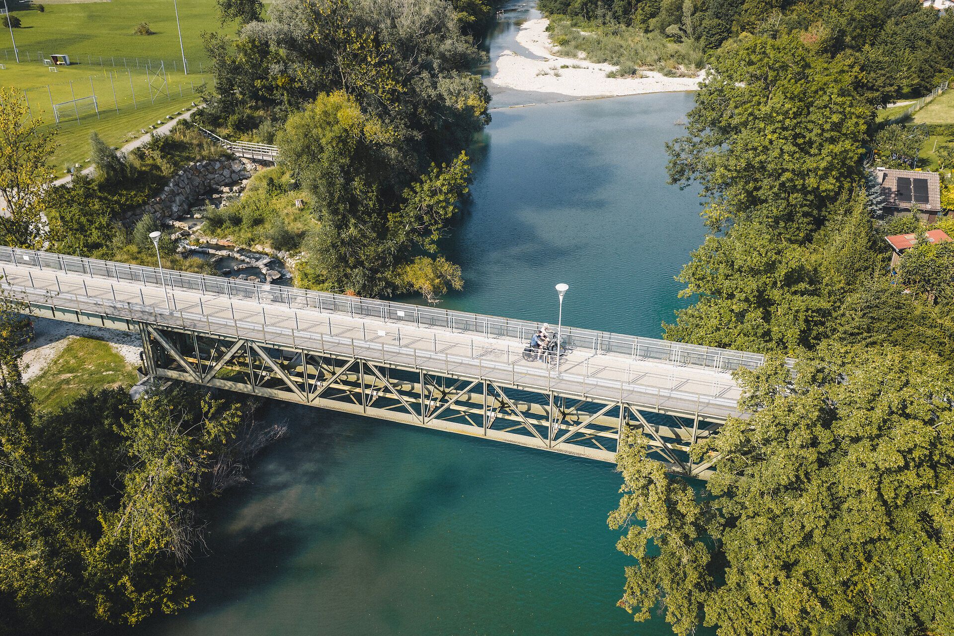 Zwei Radfahrer fahren auf dem Erlauftalradweg über eine Brücke, darunter der tiefblaue Fluss und daneben grüne Landschaft entlang des Ufers.