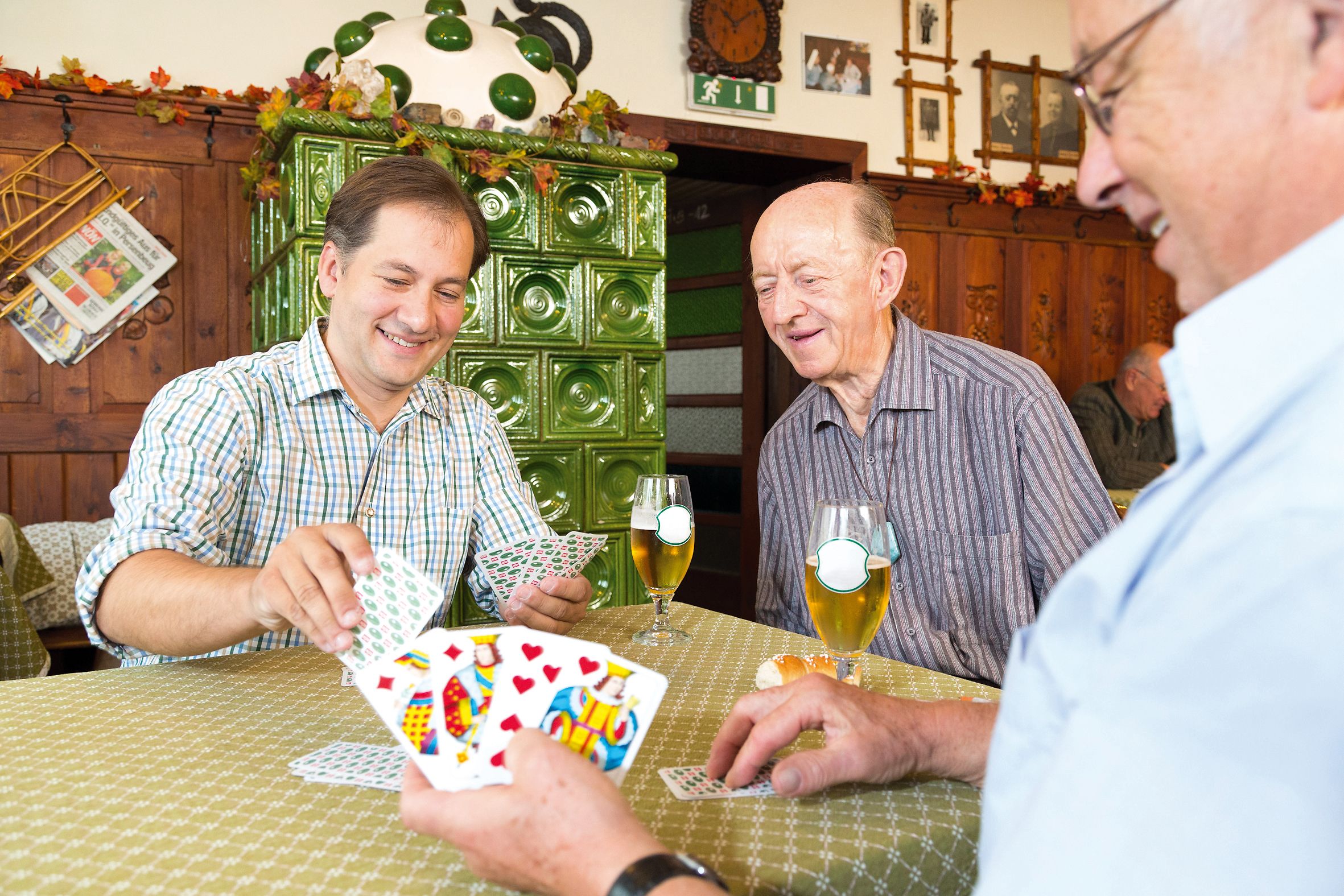 Drei Männer spielen Karten in einem Gasthof mit grünem Kachelofen im Hintergrund.