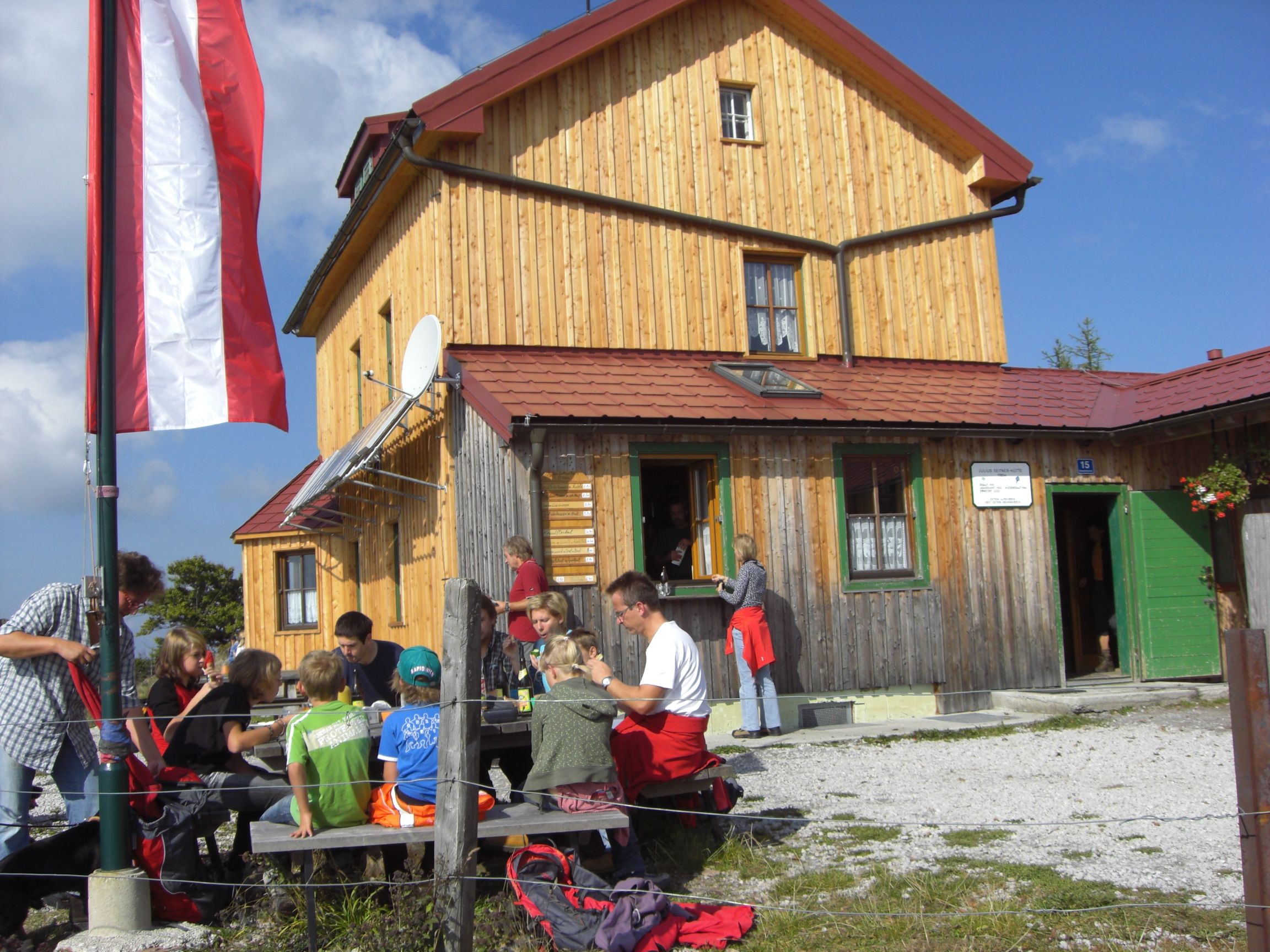 Eine Gruppe von Menschen sitzt vor einer Berghütte mit Holzfassade und österreichischer Flagge.