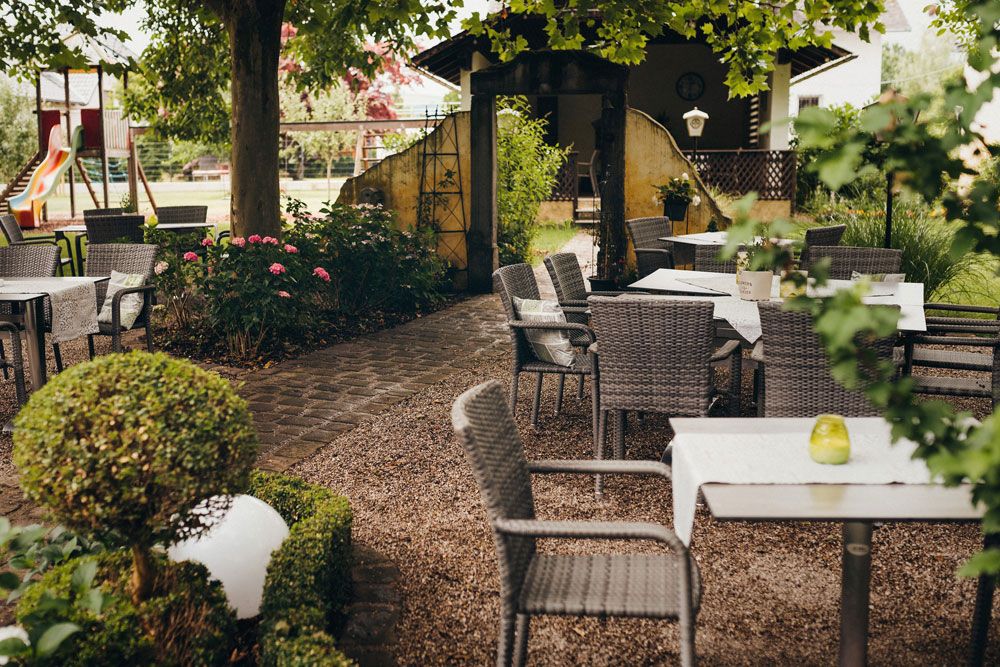 Ein gemütlicher Außenbereich mit Tischen und Stühlen unter Bäumen, umgeben von Pflanzen und einem Spielplatz im Hintergrund.